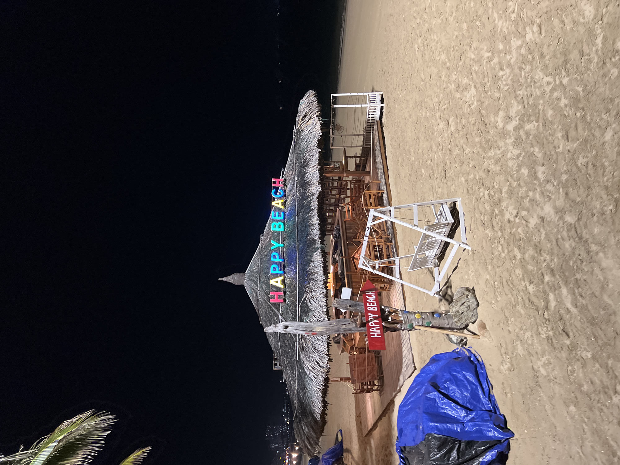 The image shows a beach bar called "HAPPY BEACH" at night in Nha Trang, Vietnam. The bar has a thatched roof and is lit by colorful neon signs. There are tables and chairs set up outside the bar, and a swing set is visible in the foreground. The beach is sandy, and the ocean is in the background. A large blue tarp covers some objects to the left of the frame. The overall scene suggests a lively but potentially quiet beach bar at night.