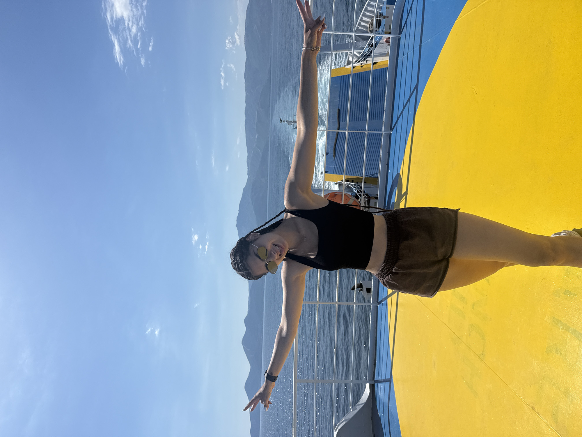 A young woman with dark braided hair, wearing sunglasses, a black tank top, and brown shorts, stands on the deck of a ferry with her arms outstretched. The deck is painted yellow and blue. In the background, the sea is visible, with a wake trailing behind the ferry, and distant mountains under a clear blue sky with scattered clouds. The scene appears to be in Greece, given the mention of the location.