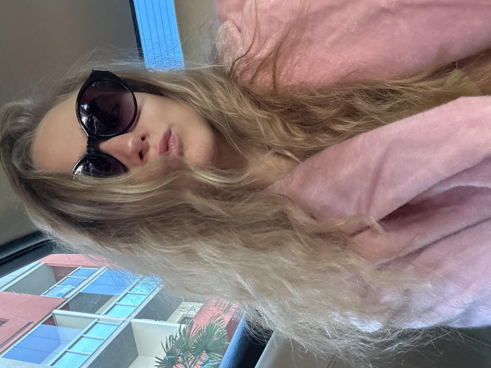 A woman with long blonde hair and sunglasses is in a car. She is wearing a pink fuzzy jacket. The buildings outside the car are peach and white with blue windows. A palm tree is visible outside the car. The location is Unknown, Unknown.