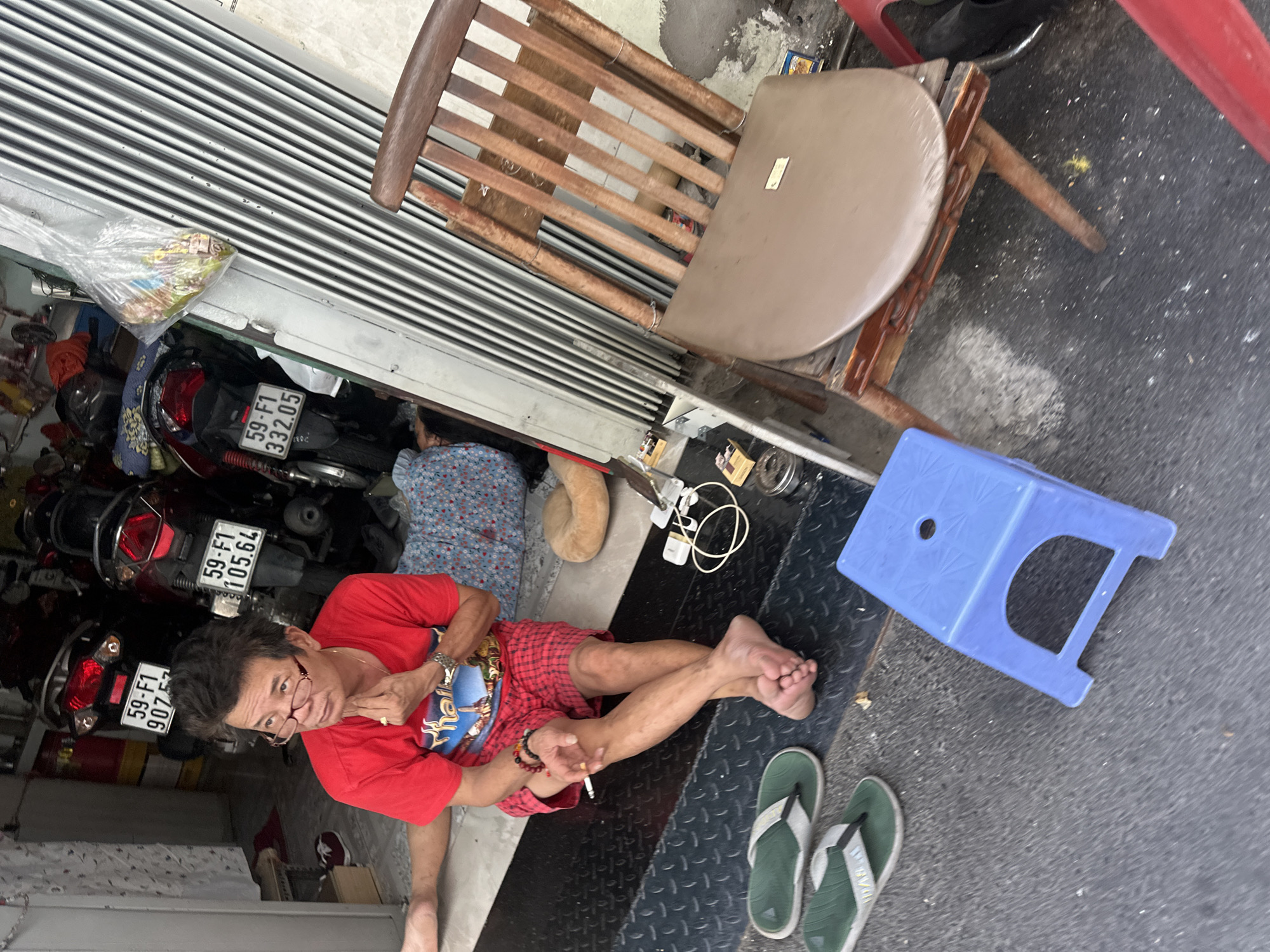 The image is taken at eye-level, looking down on an outdoor scene in Thủ Đức, Vietnam. A man wearing glasses, a red T-shirt, and red plaid shorts is sitting on the ground smoking a cigarette. He is surrounded by a metal storefront door, a wooden chair, and a blue plastic stool. Next to him are a pair of green and white flip-flops. In the background, there are two motorbikes parked with license plates that read "59-F1 907.57", "59-F1 105.64", and "59-F1 332.05". An object that appears to be a bag of snacks hangs from the storefront door. A pillow is on the ground behind the seated man.