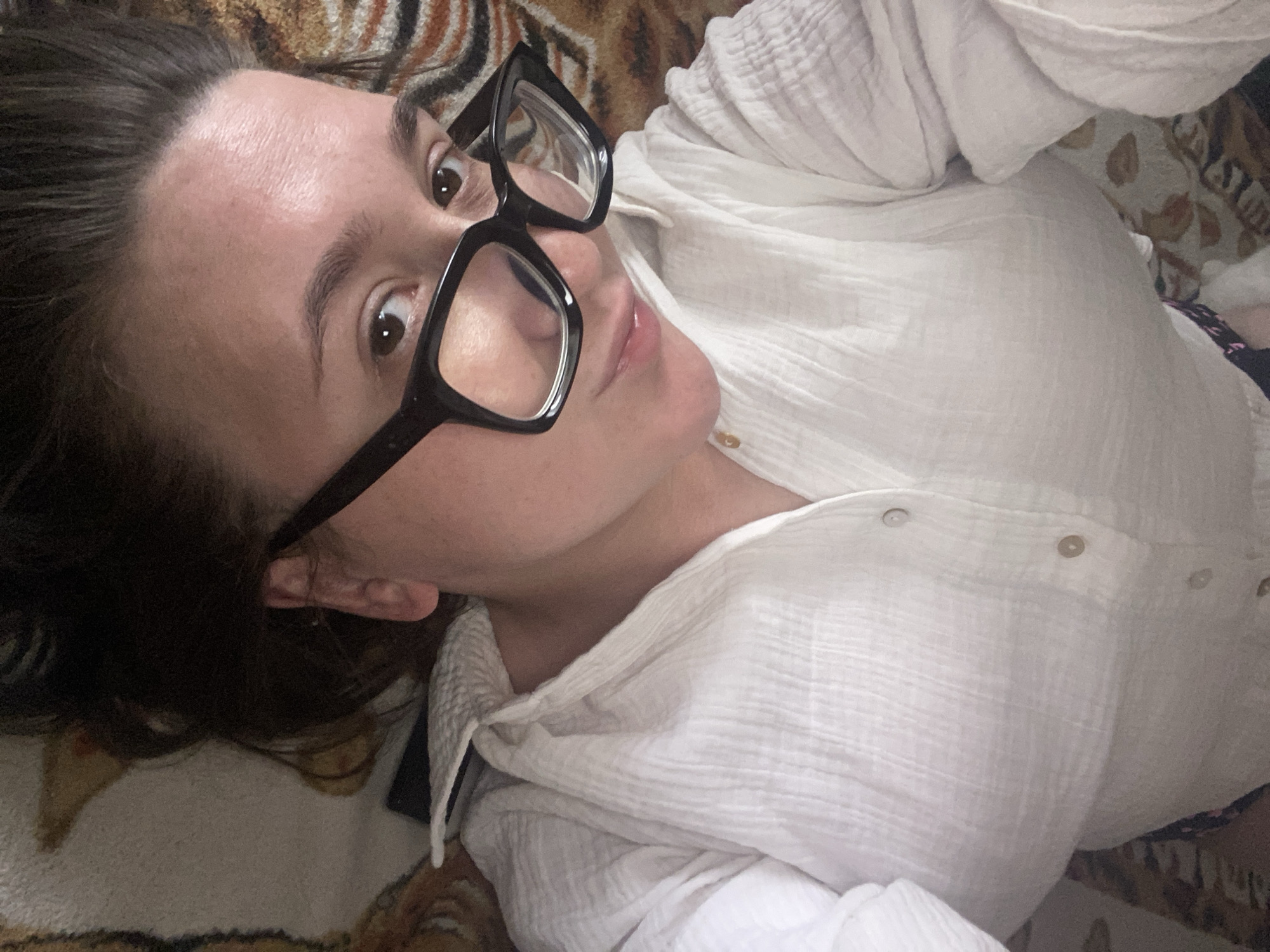 The image shows a person lying down, viewed from a high angle. The person is wearing a white, textured button-up shirt and black-rimmed glasses. Their head is tilted back, and their face is partially obscured by the glasses. The background appears to be a patterned blanket or rug. There is no discernible information in the image that would allow for the identification of a specific location in the city of Chaikovsky, Russia.