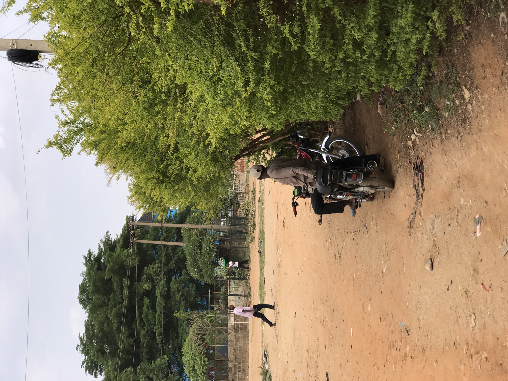The image captures a scene on a dirt road in Dutsen Alhaji, Nigeria. A man wearing a helmet and light-colored clothing is seated on a black motorcycle. Another person is also on the motorcycle, behind the driver. A person wearing a pink shirt and dark pants is walking in the opposite direction on the dirt road. Green trees line the road, and utility poles are visible in the background. A fence can be seen on the left side. The sky is overcast.