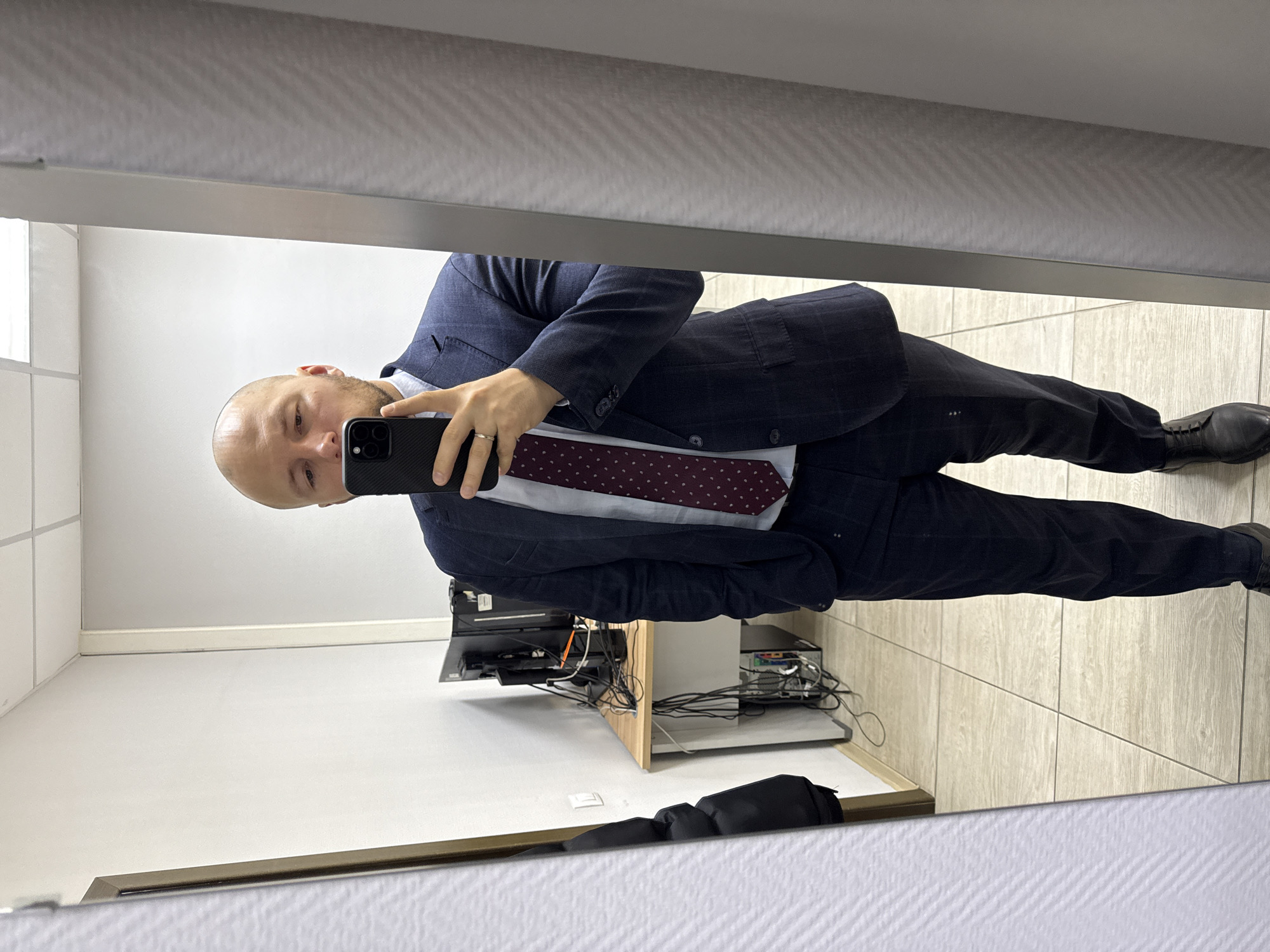 A bald man in a navy suit and maroon polka dot tie takes a mirror selfie in what appears to be an office or workspace. He holds a black smartphone. A desk cluttered with computer equipment and tangled cables is visible behind him. The flooring is light-colored tile. No specific details confirm the location as Krasnoyarsk, Russia.