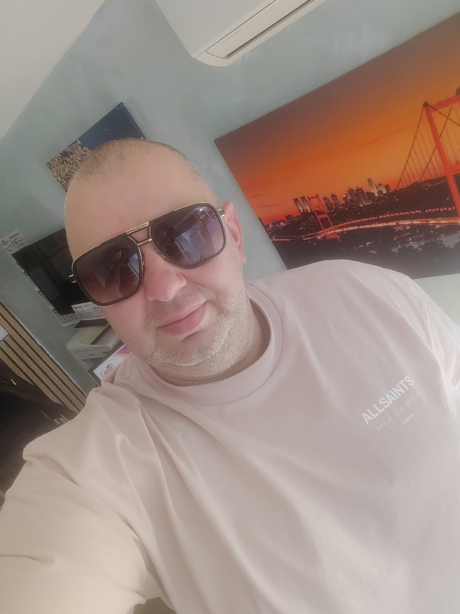 A selfie photograph shows a man wearing sunglasses and a pink t-shirt with the logo "ALLSAINTS" and Japanese text below. The background includes a cityscape print with a bridge, potentially the Bosphorus bridge in Istanbul, and another framed picture on the wall. There's an air conditioner unit mounted high on the wall. The photograph appears to have been taken indoors. I do not see visual evidence indicating that the photo was taken in Gorna Oryahovitsa, Bulgaria.