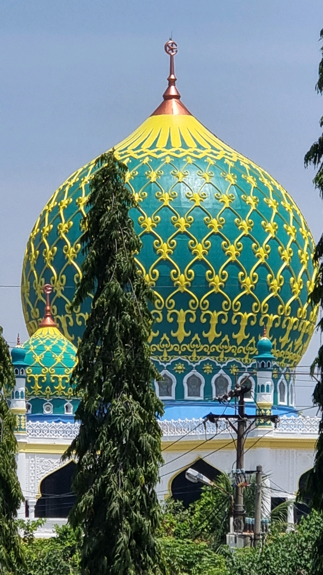 The image shows a close-up of a mosque's dome in Lhokseumawe, Indonesia. The dome is large and elaborately decorated with a teal and gold pattern. A smaller, similar dome is visible in the background to the left. Tall, green evergreen trees obscure parts of the mosque's lower structure and foreground. A utility pole with electrical wires and a street light is also visible. The sky is clear and blue.