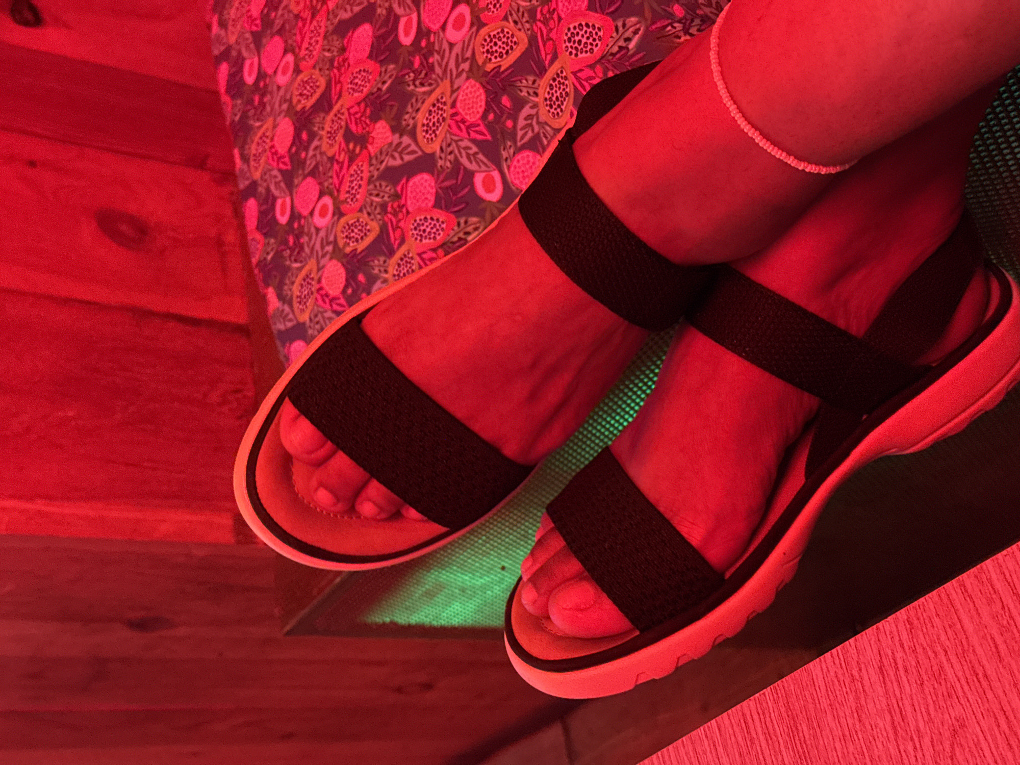 In Barcelona, Spain, a close-up shot captures a person's feet wearing black strap sandals, illuminated by intense red lighting. The sandals have thick, white soles and black elastic straps that cross over the top of the feet. One foot is positioned in front of the other, and a delicate, beaded anklet is visible on the lower leg of the back foot. The background features a wooden wall and a patterned fabric, possibly a cushion or upholstery, with pink and orange fruit designs. A patch of green light emanates from beneath the sandals, creating a contrasting element. The overall scene suggests a relaxed or recreational setting, possibly a spa or a lounge area, with the vibrant red light adding a dramatic effect.
