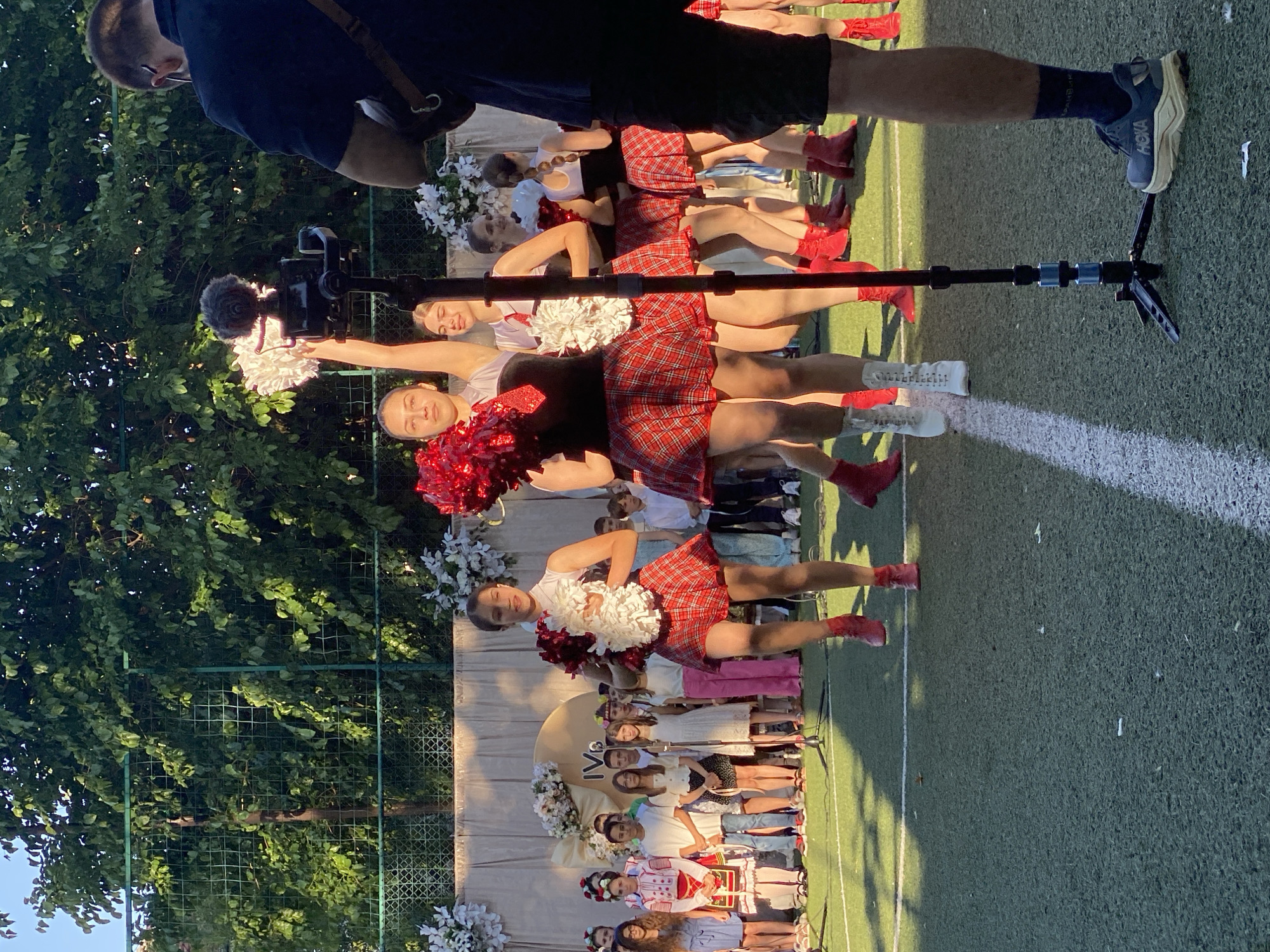 In Pleven, Bulgaria, a group of young girls in red and white cheerleader outfits perform on a green turf field. They hold pom-poms and wear plaid skirts and red boots. One cheerleader in the foreground is captured mid-jump. Behind them, a row of children in various attire, some in traditional Bulgarian clothing, watch from a stage decorated with flowers and a large, circular emblem. A cameraman stands to the right, filming the performance with a professional camera on a tripod. The scene is outdoors, with lush green trees and a clear sky in the background, suggesting a sunny day.