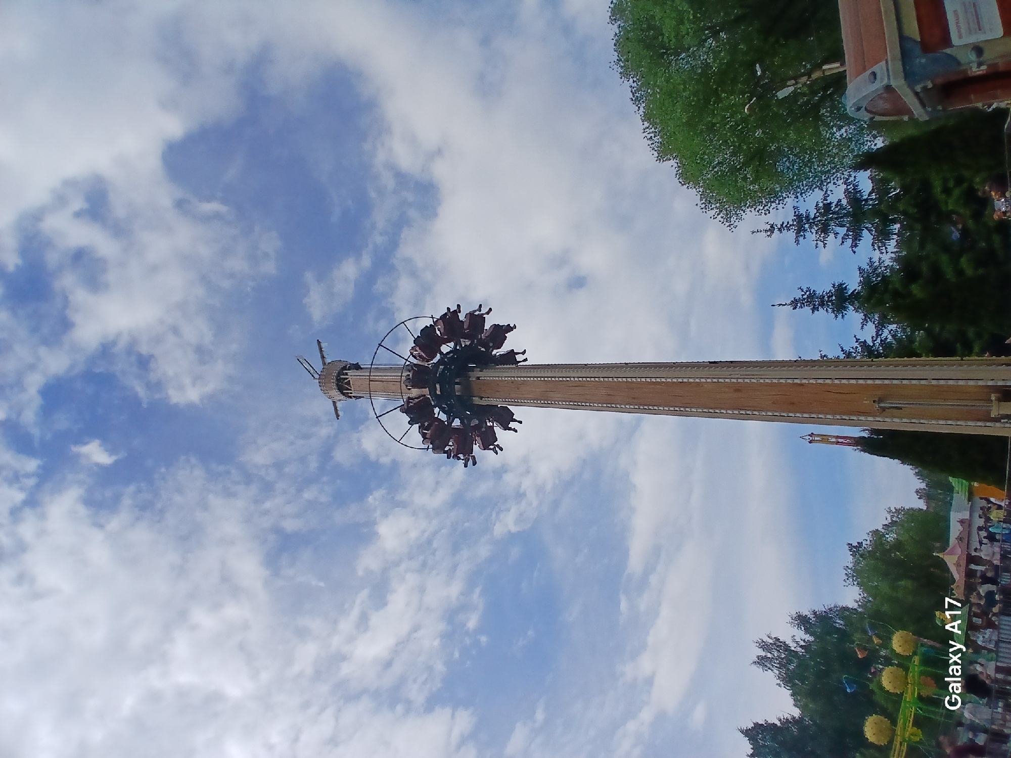 The image shows a high swing ride in the Mayakovsky Park in Yekaterinburg, Russia. The ride features a tall central tower with a circular platform at the top. Passengers are seated in swing chairs attached to the platform. The ride is in operation, with the swing chairs occupied. The sky is visible behind the ride, with blue sky and white clouds. Trees are seen in the background around the ride. A Galaxy A17 watermark is visible in the corner of the image.
