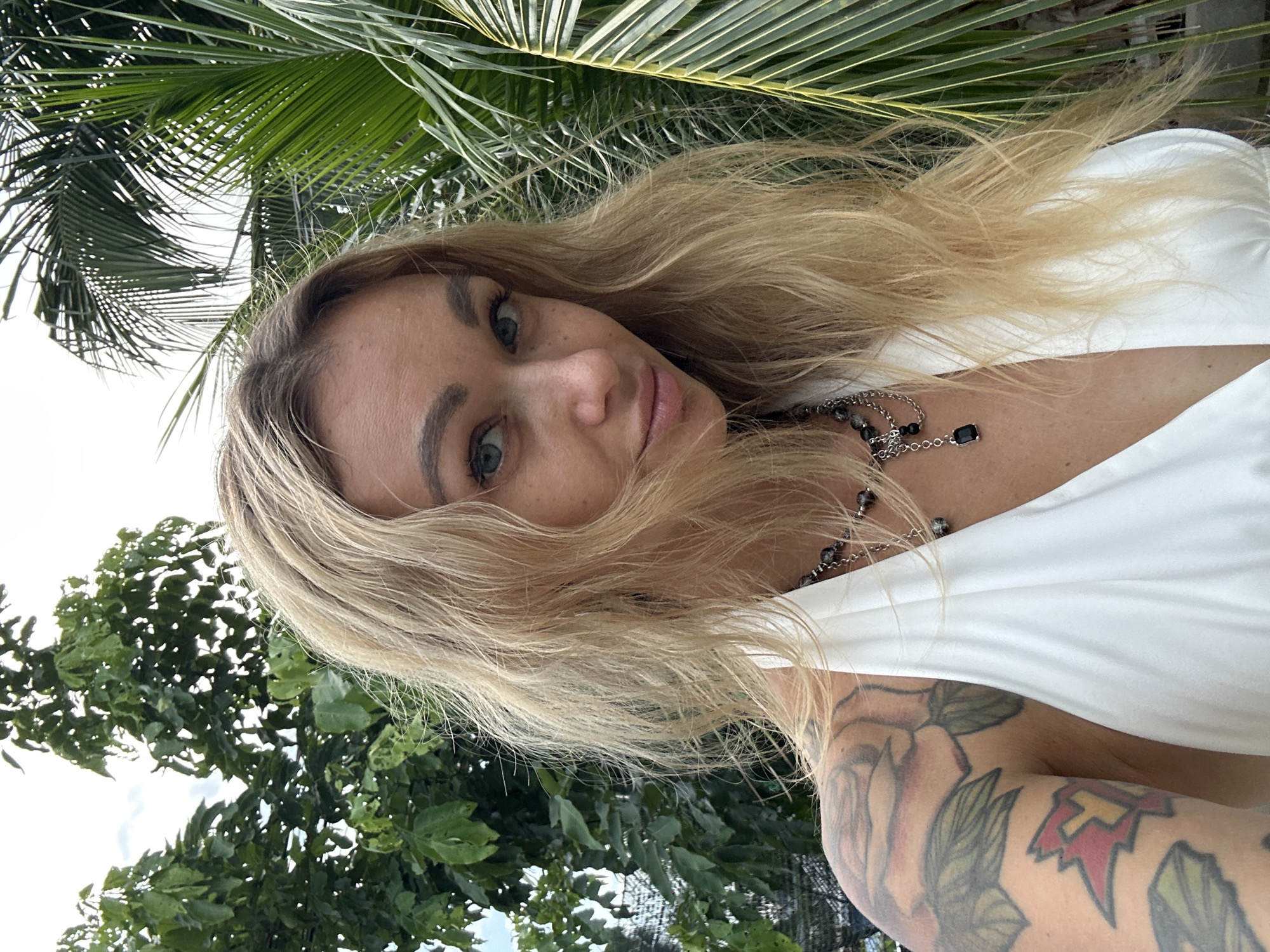 A woman with fair skin and blonde hair poses for a selfie outdoors in Mũi Né, Vietnam. She is wearing a white top and a silver necklace with black stones. Her right arm, visible in the foreground, is heavily tattooed. The background features lush green foliage, including palm trees. The lighting suggests daytime.