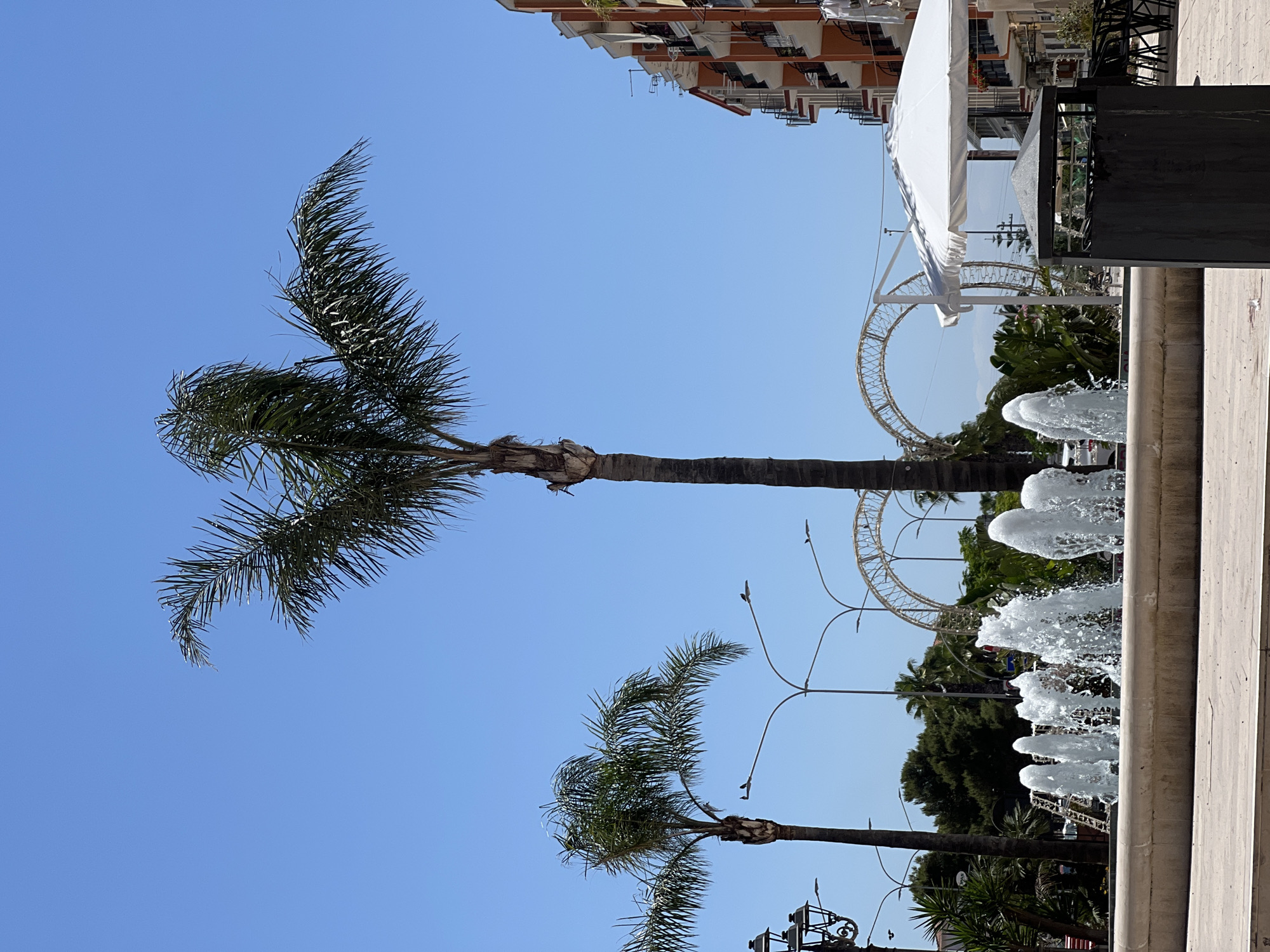 The image captures a street scene in Margherita di Savoia, Italy. In the foreground, a row of water fountains spray water into the air from a stone basin. To the right of the fountains, a dark trash receptacle stands next to outdoor seating with chairs and tables, covered by a white umbrella. Further back, two tall palm trees frame the scene, with one centrally positioned and another to the left. In the mid-ground, a street lamp with multiple lights is visible on the left, and in the background, a decorative arch structure can be seen. A multi-story building with balconies and orange and white facade is on the right side of the image. The sky is clear and blue, indicating a sunny day. No people are visible in the image.