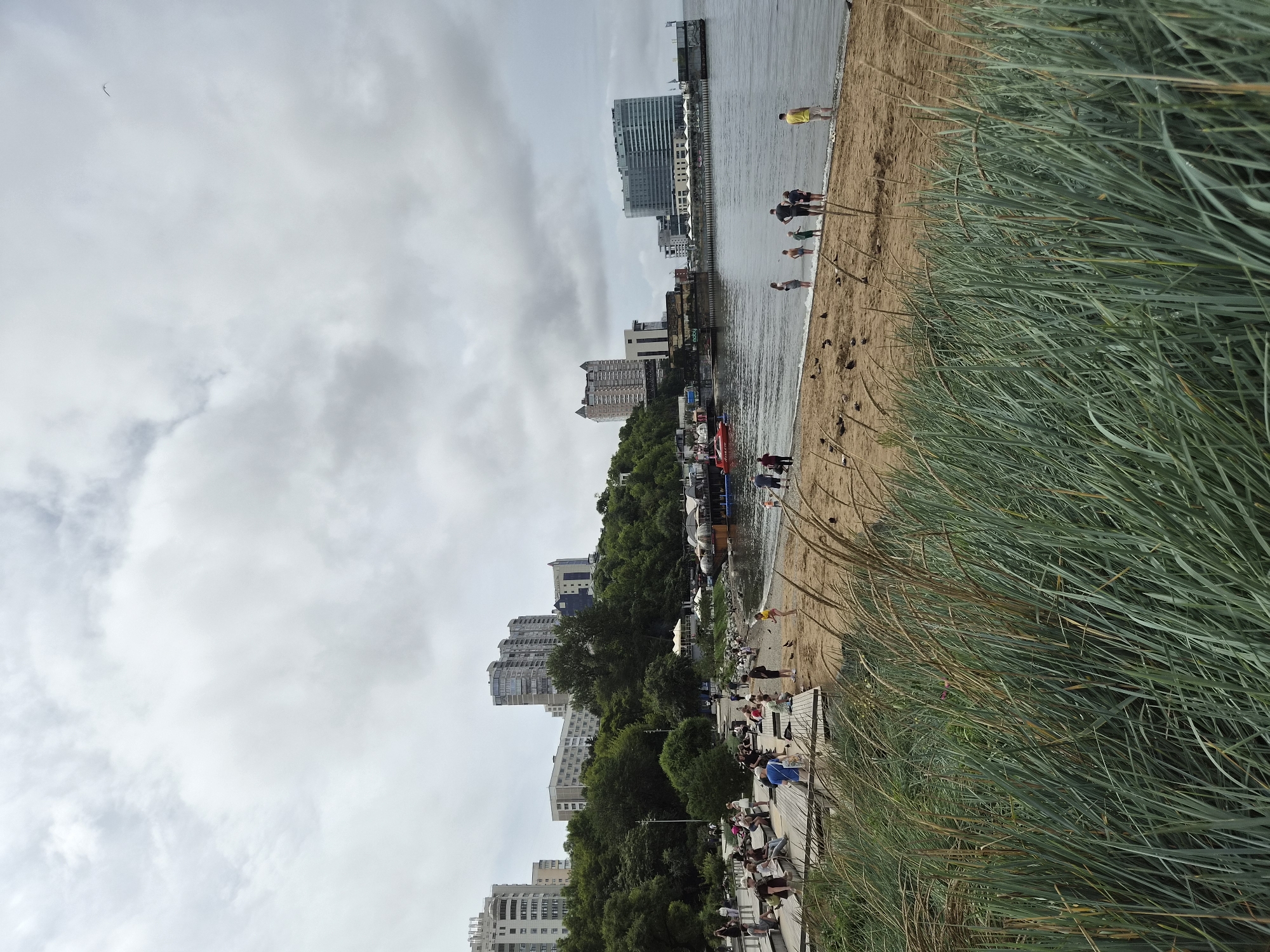 The image depicts a cloudy day at a beach in Vladivostok, Russia, specifically at the Zolotoy Rog Bay. In the foreground, tall green grass with dried stalks blows in the wind. Behind the grass, a sandy beach is populated with several individuals and groups. Some people are seated on a wooden boardwalk structure, while others are standing or walking on the beach. Several people are near the water's edge, with a few children wading in the shallow surf. A family of four, including two adults and two children, are standing in the water. Further down the beach, a lone individual in a yellow shirt stands near the water. Several small birds are scattered on the sand.
The middle ground features the calm, grey water of the bay. Moored boats, including a distinctive red boat and several restaurant-style vessels, are visible near the shoreline. The background is dominated by a densely wooded hillside, characteristic of Vladivostok's topography. On top of the hill, several modern, multi-story buildings and hotels are visible, including a large, glass-fronted hotel on the far right. The sky is overcast with varying shades of grey and white clouds.