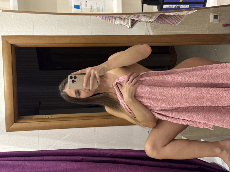 In a bathroom mirror selfie, a woman holds a phone with her right hand, partially obscuring her face. Her long, straight hair cascades over her shoulder. She's wrapped in a textured, light pink towel. The bathroom features white tiled walls, a wooden-framed mirror, and shelves with various toiletries above. A purple curtain is visible at the bottom of the frame.
