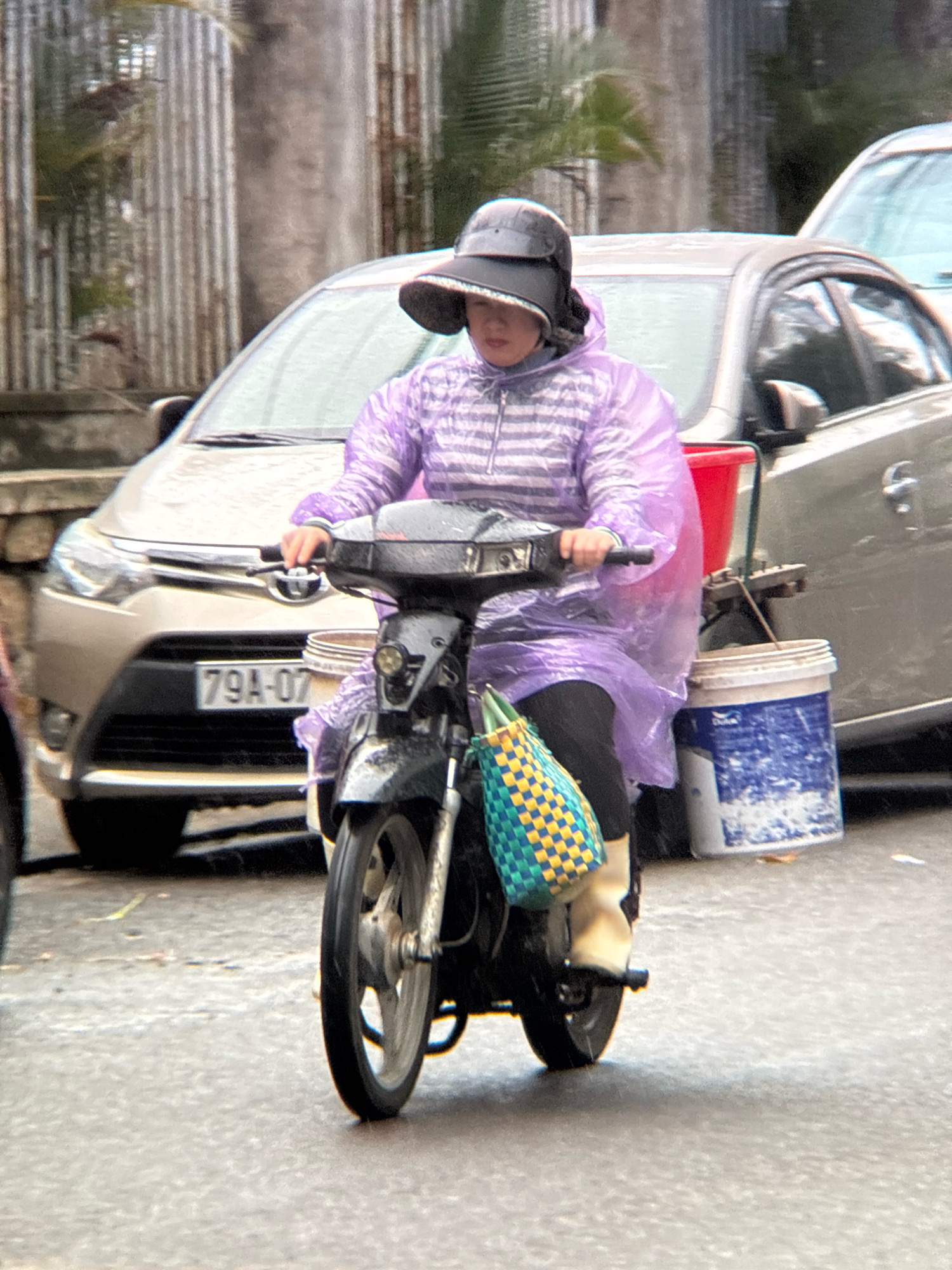In Nha Trang, Vietnam, a person wearing a purple striped hooded jacket and a wide-brimmed hat rides a black scooter on a wet asphalt road. The person's face is partially obscured by the hat. The scooter is equipped with a green and yellow checkered bag hanging from the front and two buckets attached to the rear, one red and one blue and white. In the background, a silver car with a license plate reading "79A-07" is visible, along with a corrugated metal fence and foliage. The lighting suggests an overcast or rainy day.