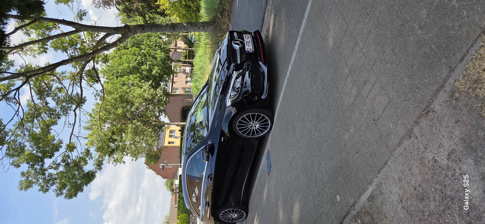 A black Mercedes-Benz CLA Shooting Brake station wagon is parked on the side of a paved road in Eeklo, Belgium. The car is facing right, with its front end slightly turned towards the viewer. The license plate reads "2-FBL-620". The car is parked on the right side of the road, with a sidewalk to its left. The background consists of residential buildings, trees, and green grass. The sky is blue with scattered white clouds. A traffic signal is visible in the distance. The image is captured from a slightly low angle, emphasizing the car. The lower left corner of the image shows a "Galaxy S25" watermark.