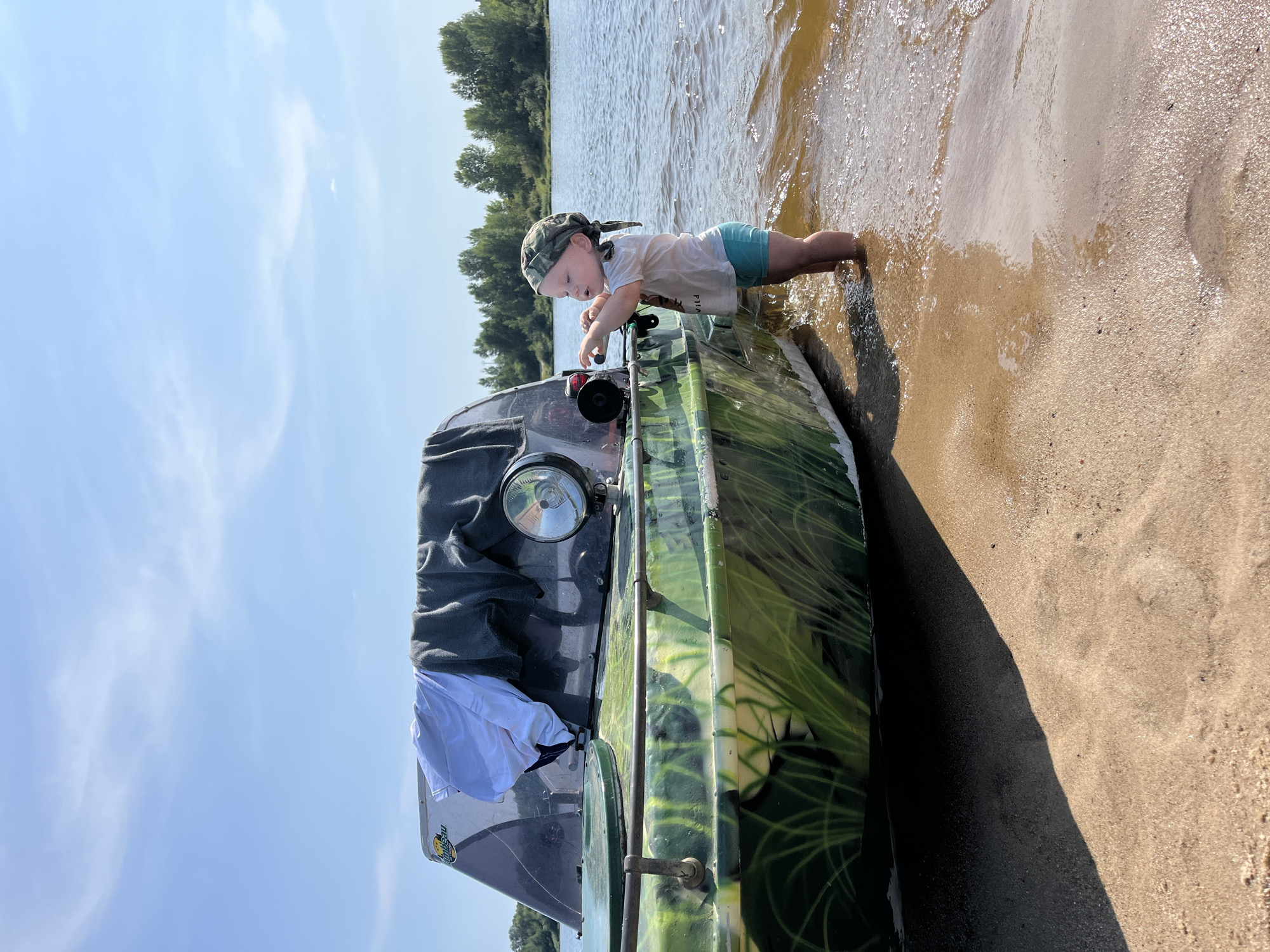 A child stands in shallow water next to a camouflage-painted boat on a sandy shore. The boat has a windshield and a large spotlight. Clothes are draped over the windshield. The child is wearing a camouflage bandana, a white t-shirt, and blue shorts. Trees line the opposite bank of the river in the background. The sky is blue with wispy clouds. The location is a riverbank in Nizhny Novgorod, Russia.