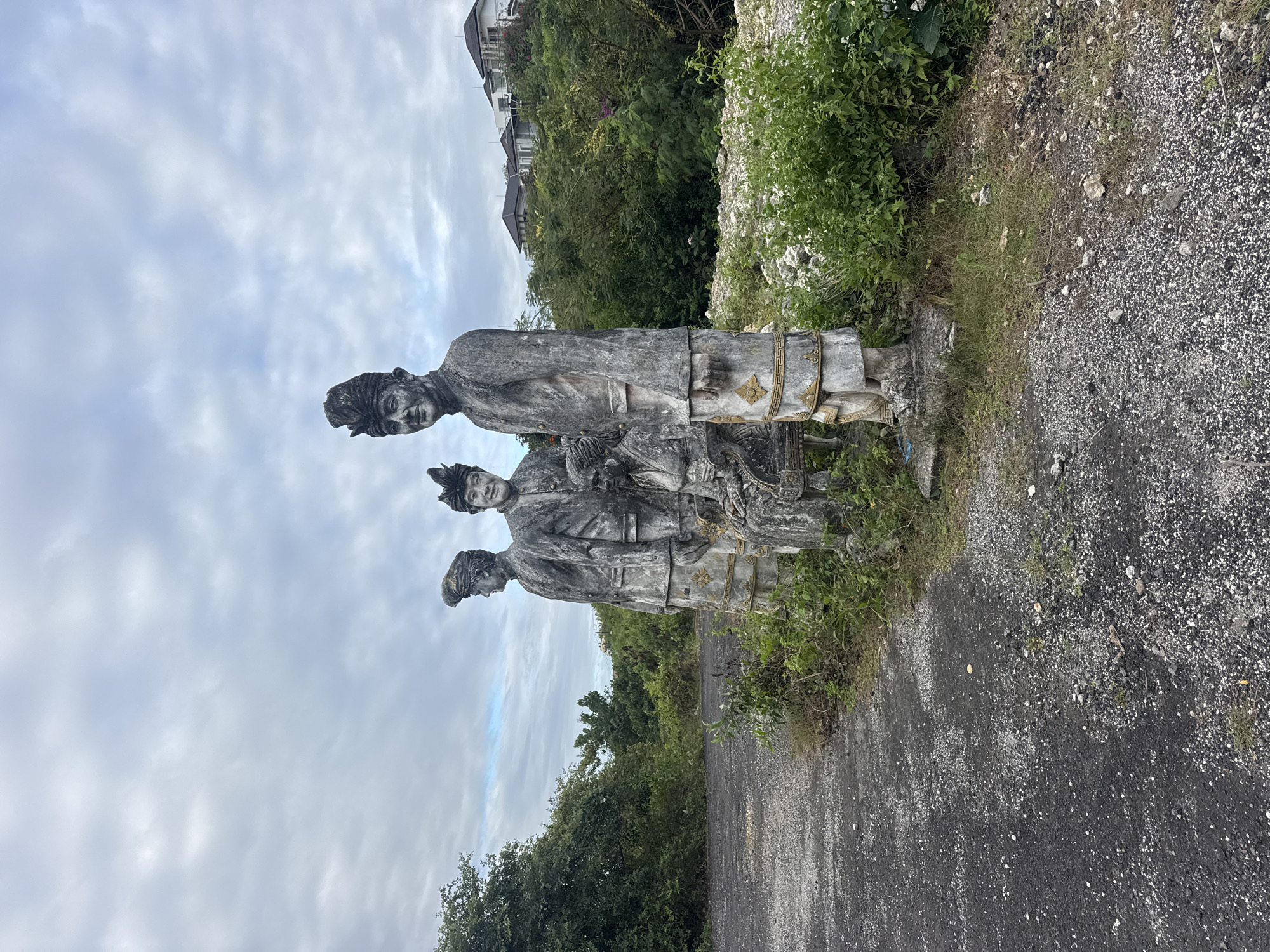 In Kuta Selatan, Indonesia, a series of weathered concrete statues stands on an overgrown, unpaved lot. The statues depict three individuals in traditional attire, possibly ceremonial clothing, with the two prominent figures on the right appearing to be men wearing ornate hats and long jackets. A third, smaller figure is seated between them. The statues are positioned near a paved, rough road and surrounded by lush green vegetation and scattered white stones. In the background, modern houses are visible on a hillside under a cloudy sky. The scene suggests a potentially abandoned or developing site, with the statues serving as a historical or cultural marker.