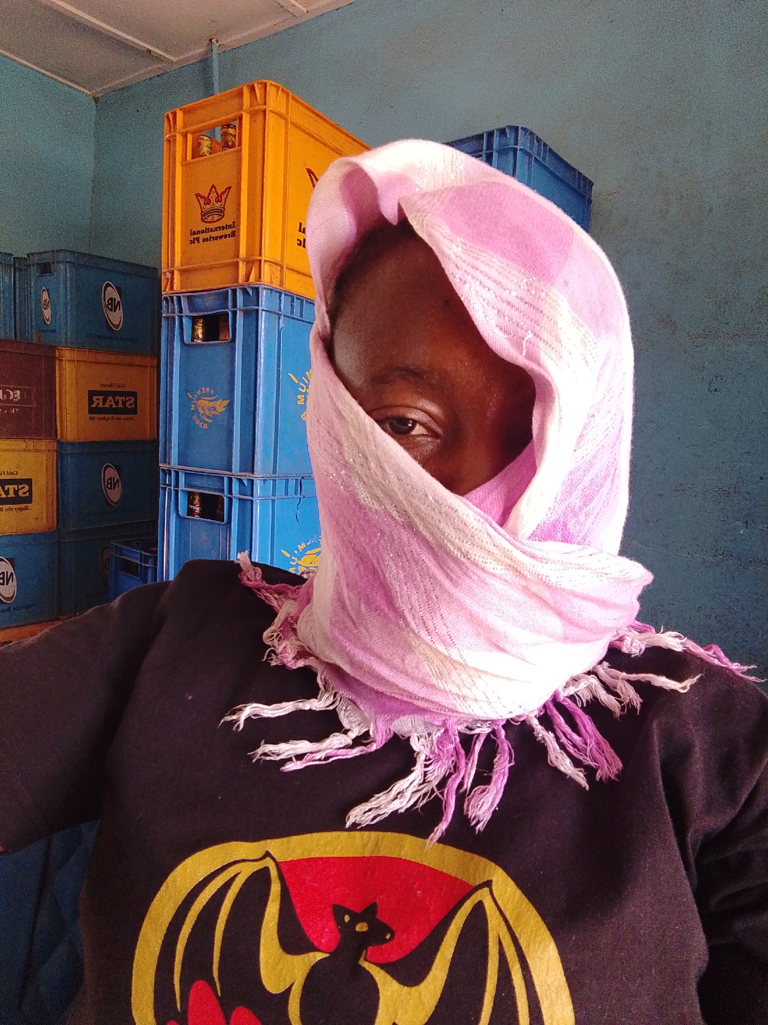 A person stands indoors, their face mostly concealed by a pink and white scarf with fringed edges. Only one eye is visible. The person wears a black long-sleeved shirt with a large, colorful emblem that appears to feature a bat. In the background are stacks of blue, yellow, and brown plastic crates, likely used for beverages, with filled glass bottles visible inside some. The wall is a faded blue. The location is in the city of Unknown, Unknown.