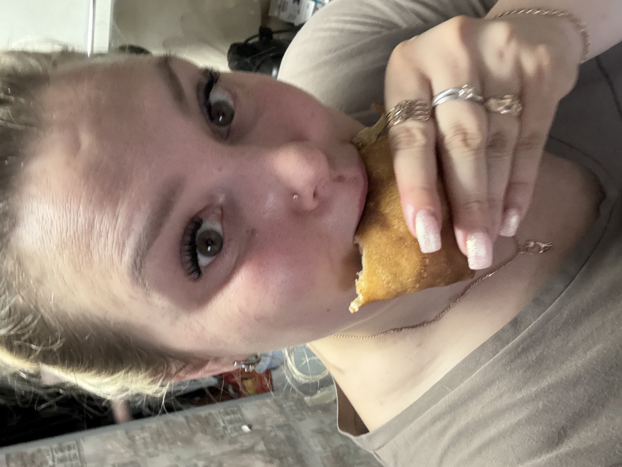 A close-up, tilted selfie shows a woman with fair skin, blond hair, and green eyes eating something. She has a nose piercing and is wearing multiple rings on her fingers and a delicate gold necklace. The background is blurred and out of focus. The woman is wearing a tan colored shirt. The image does not provide sufficient information to identify the location as Марковский, Russia.