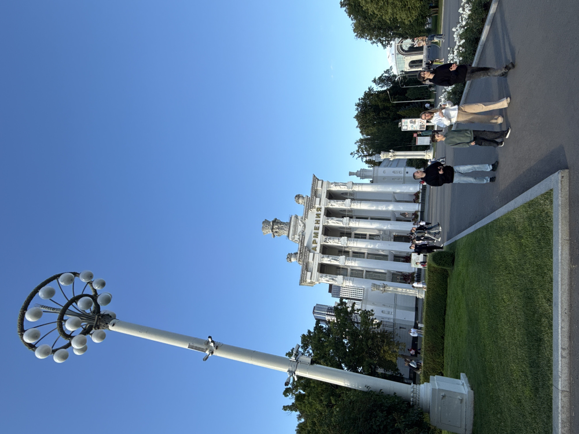 The image depicts the architectural complex of VDNKh (Exhibition of Achievements of National Economy) in Moscow, Russia. In the foreground, a tall, white lamppost with a circular, spiral arrangement of light bulbs stands on the left. The lamppost has several security cameras attached. To the right, a family of four, consisting of two adults and two children, is walking on a paved path. The father, wearing a black hooded sweatshirt and blue jeans, is in the lead, followed by a young boy in a green hooded sweatshirt and black pants. Behind them, a woman in a white hooded sweatshirt and light-colored pants is walking, and a teenage boy in a black hooded sweatshirt and dark jeans is on the far right of the group.
In the background, the grand white building of the "Armenian SSR" pavilion at VDNKh is visible, characterized by its classical architecture, numerous columns, and ornate sculptures. The pavilion has the word "АРМЕНИЯ" (Armenia) inscribed above its entrance. Surrounding the pavilion are manicured green lawns, hedges, and mature trees. The sky is a clear, bright blue. In the distance, other VDNKh structures and modern buildings are visible. The scene suggests a pleasant day at a public exhibition or park.