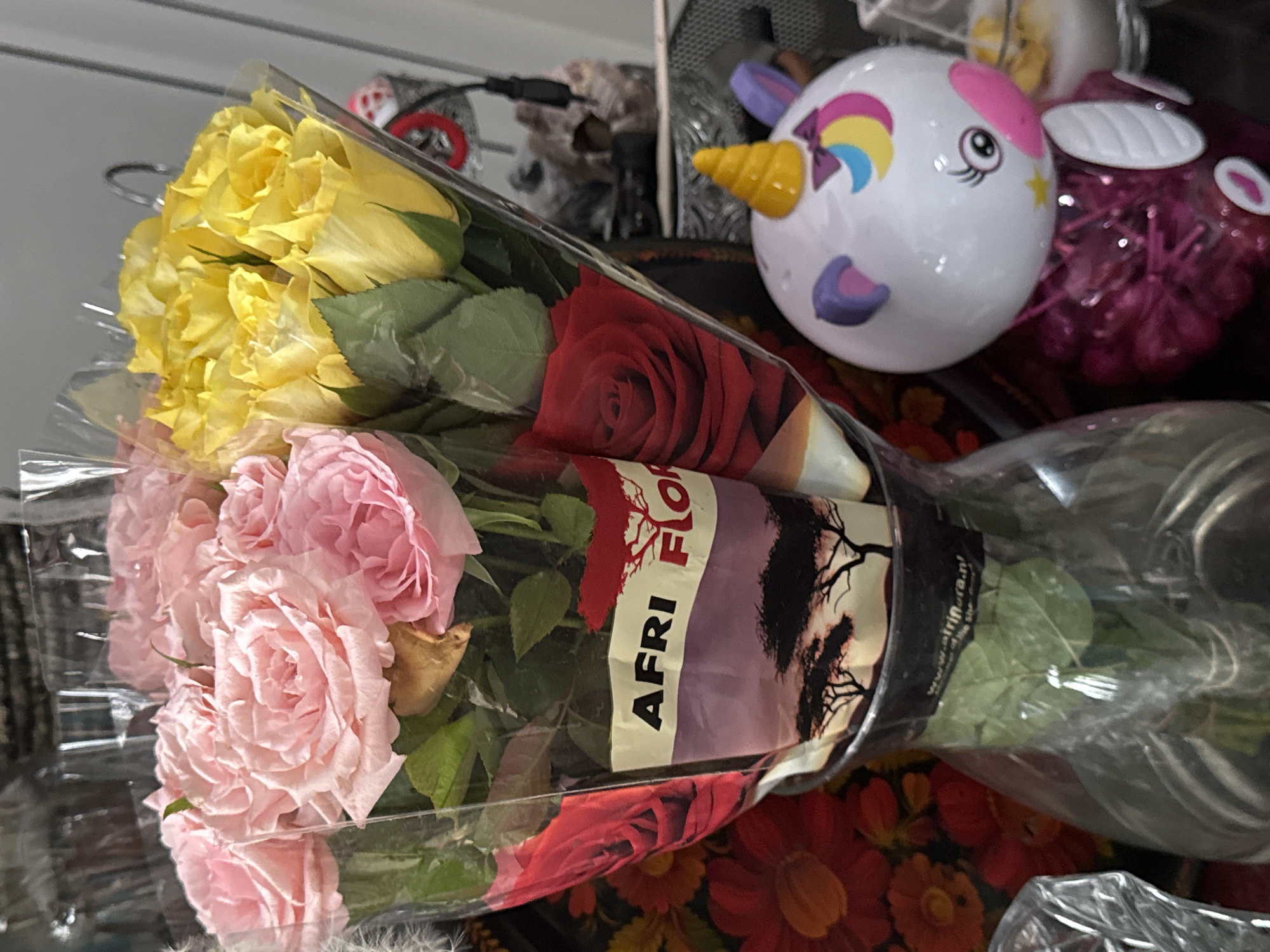 The image shows a bouquet of pink and yellow roses in a glass vase. The flowers are partially wrapped in plastic packaging with text that appears to be "AFRI FLOR". There is also a decorative item resembling a unicorn with a yellow horn and colorful accents to the right of the vase. In the background, there are other objects and decorations, including what appears to be a dark metal rack and a red floral arrangement. The scene is dimly lit. The image is a photograph taken indoors, likely in a home environment. No specific location in the city of Hallum, Netherlands is depicted or suggested by the visible elements in the image.