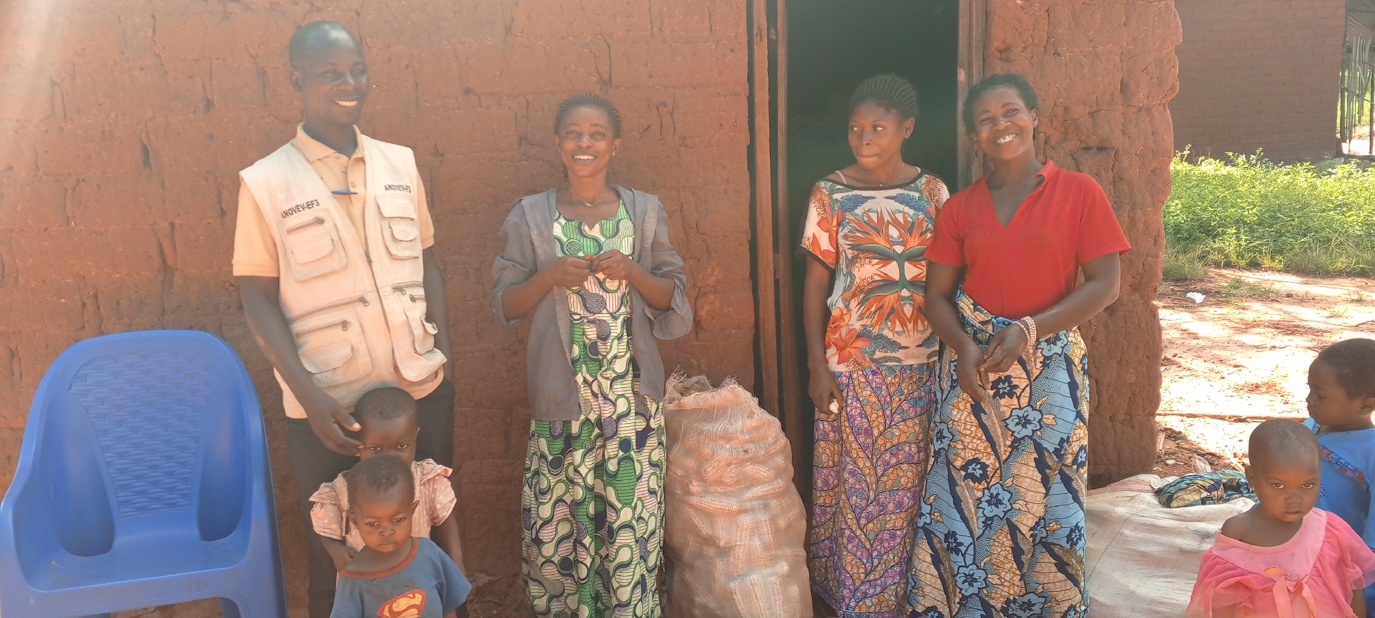 Outside a mud brick building in Ngalula, Democratic Republic of the Congo, four women and three young children are positioned in front of the building's entrance and adjacent wall. A man wearing a khaki-colored vest stands to the left of the group. To his right stands a woman wearing a long, green patterned dress, a gray sweater, and a silver necklace. Next to her is a burlap sack of potatoes. Another woman in an orange floral top and long, purple patterned skirt is partially obscured by the potatoes. Finally, a woman in a red shirt and long, blue patterned skirt stands on the right side of the group. Two children stand on the left, partially obscured by a blue plastic chair, and two other children stand to the right.