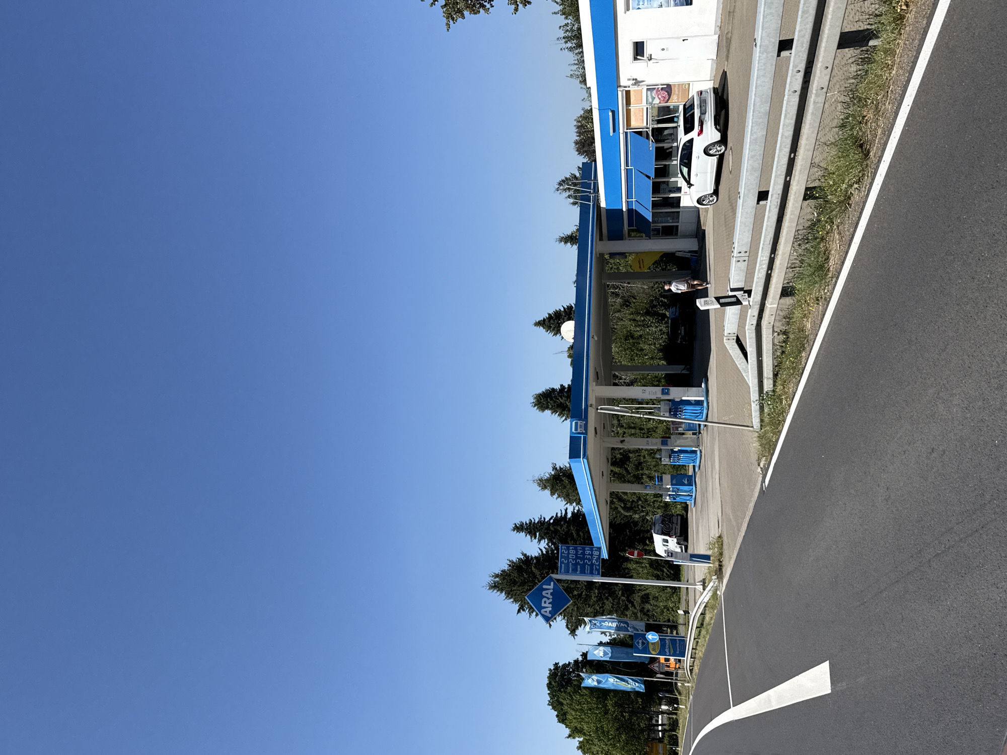 The image depicts a gas station in Herzberg, Germany, on a clear, sunny day with a vibrant blue sky. In the foreground, a paved road curves into the frame, marked by a white line. A metal guardrail separates the road from a grassy embankment.
The central focus is the ARAL gas station, characterized by its bright blue canopy and white building. Several gas pumps are visible under the canopy, some with blue accents. A white Audi station wagon is parked in front of the building. A person is walking near the pumps, and another person is visible by the building entrance.
A prominent blue ARAL sign stands to the left of the gas station, with a price board displaying fuel costs. Several blue flags with the ARAL logo are also displayed. Behind the gas station, a line of mature evergreen and deciduous trees creates a natural backdrop. A white camper van is parked near the gas pumps. The overall scene conveys a typical, functional environment of a roadside service station.