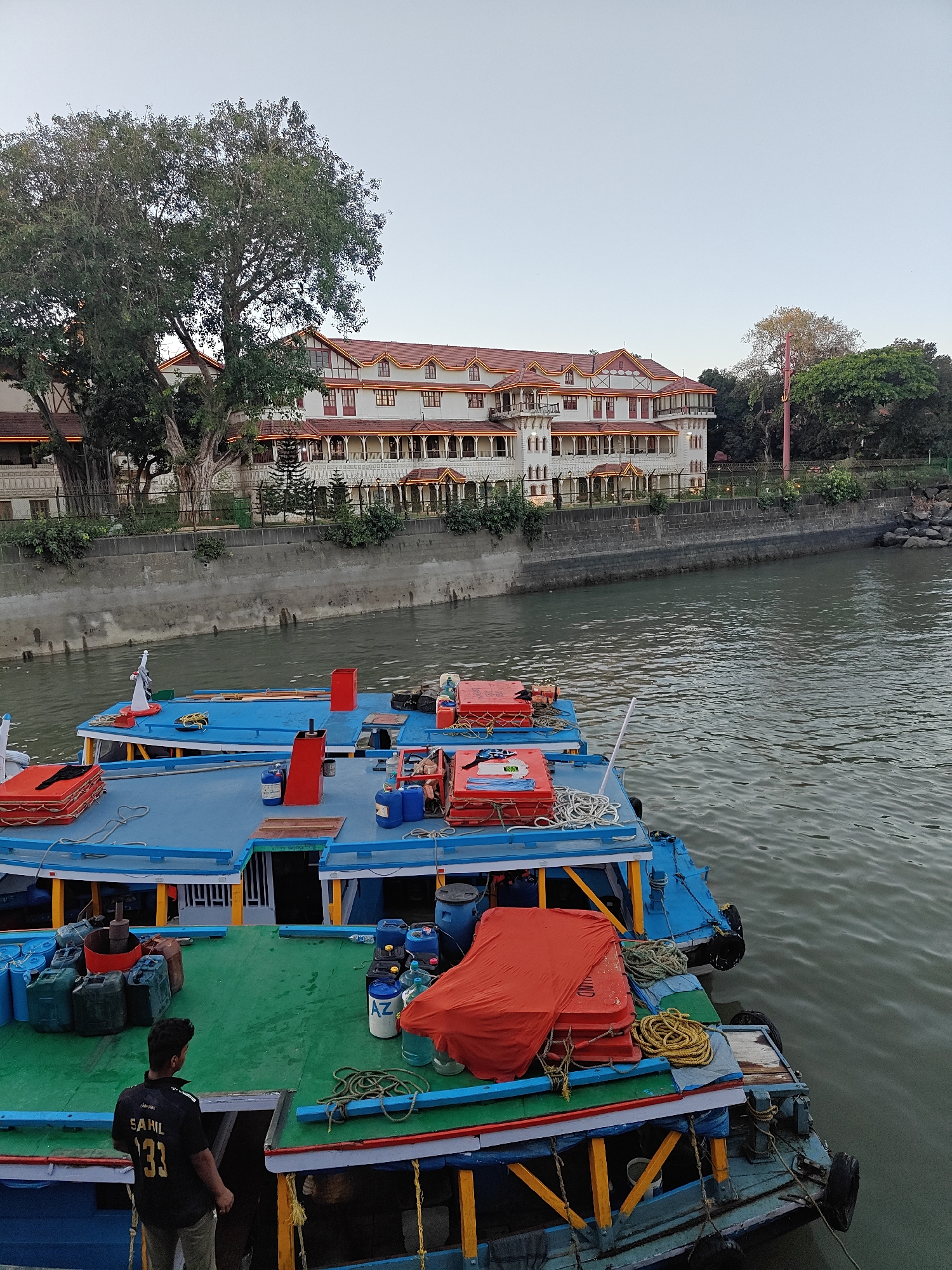 The image depicts a waterfront scene in an unidentified city. In the foreground are several boats docked closely together, their decks painted in blue, green, and yellow. Various objects clutter the decks, including red safety equipment, blue barrels, containers, and coils of rope. A man wearing a dark shirt with the name "SAHIL" and the number "33" on the back stands on one of the boats, facing away from the camera.
Beyond the boats, a concrete embankment lines the water's edge. Behind the embankment stands a large, multi-story building with a white facade and red tiled roof. The building is decorated with ornamental details and lights. Trees surround the building, including a large banyan tree on the left side. The sky is a pale blue, suggesting either early morning or late afternoon. The water in the foreground appears calm and greenish-grey.