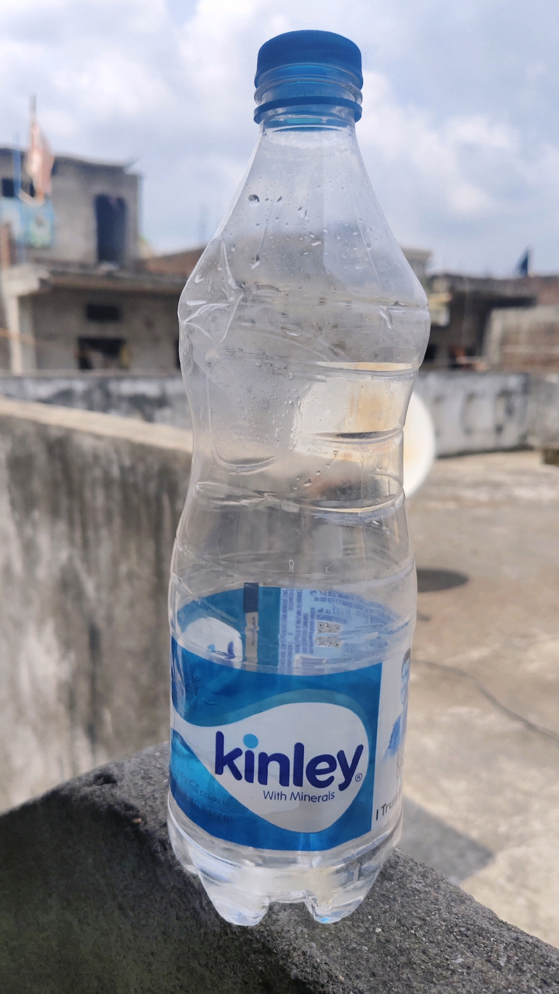 An eye-level, medium shot captures a partially full, clear plastic bottle of Kinley water. The bottle, positioned in the foreground, is resting on a dark, rough-textured surface that appears to be a ledge or wall. Condensation is visible on the upper portion of the bottle. The bottle's label, primarily white and blue, prominently features the "kinley" logo in blue text and the words "With Minerals" below it.
In the background, out of focus, are the rooftops and structures of buildings, suggesting an urban or semi-urban environment. The sky above is partly cloudy, with a mixture of blue sky and white clouds. The overall impression is that of a simple, everyday scene set outdoors.
While the image itself does not contain explicit indicators of Thikri, India, if this location is indeed Thikri, then the scene depicts an outdoor setting within that specific Indian locale, characterized by typical Indian architecture and common consumer products like bottled water.