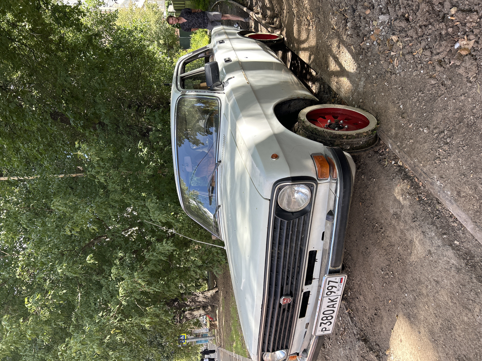 A light-colored sedan is parked on a dirt road next to a sidewalk lined with trees. The car has a license plate that reads "P380AK 997 RUS". A young man wearing a patterned shirt and gray shorts stands to the right of the car, looking at the camera. In the background, pedestrians can be seen walking on the sidewalk, and buildings are visible through the trees. The scene appears to be an urban environment on a sunny day.
