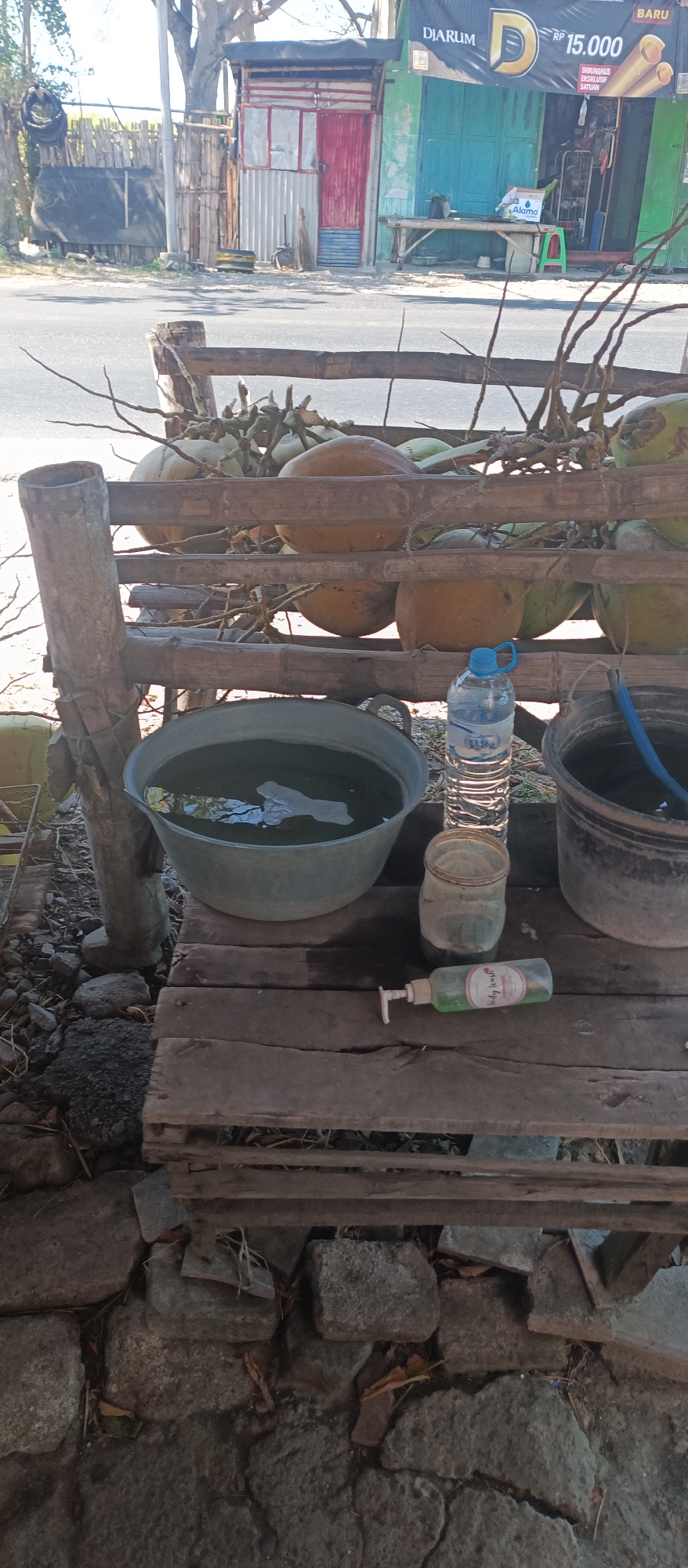 The image depicts an outdoor scene with a focus on a rustic wooden rack displaying several coconuts. Beneath the rack, a large, shallow basin filled with water sits on a wooden platform, next to a black bucket with a blue hose. A plastic bottle of water, a glass jar, and a green pump bottle labeled "Body Wash" are also present on the platform. The background reveals a roadside stall with a corrugated metal structure, a blue-painted facade, and a banner with Indonesian text and pricing. The environment suggests a commercial setting, possibly a roadside vendor in Jatisari, Indonesia, given the visual cues of the stall and the coconuts. The ground is covered with stones and debris, indicating an unpaved area. The lighting suggests daylight, with a slightly overcast sky.