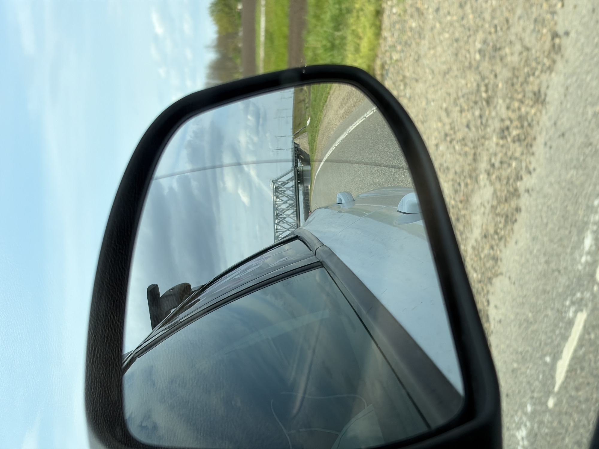 The image depicts a view from a car's side mirror, showcasing a gray bridge with a metal framework reflected in the mirror. The mirror's frame is black, and the side of the vehicle is visible within the mirror's reflection. Outside the mirror, the road is visible on the right, adjacent to the edge of the mirror. Beyond the road in the mirror's reflection, there is a stretch of green landscape. The sky is overcast. It can not be confirmed that this image is located in Medvyodovskaya, Russia.