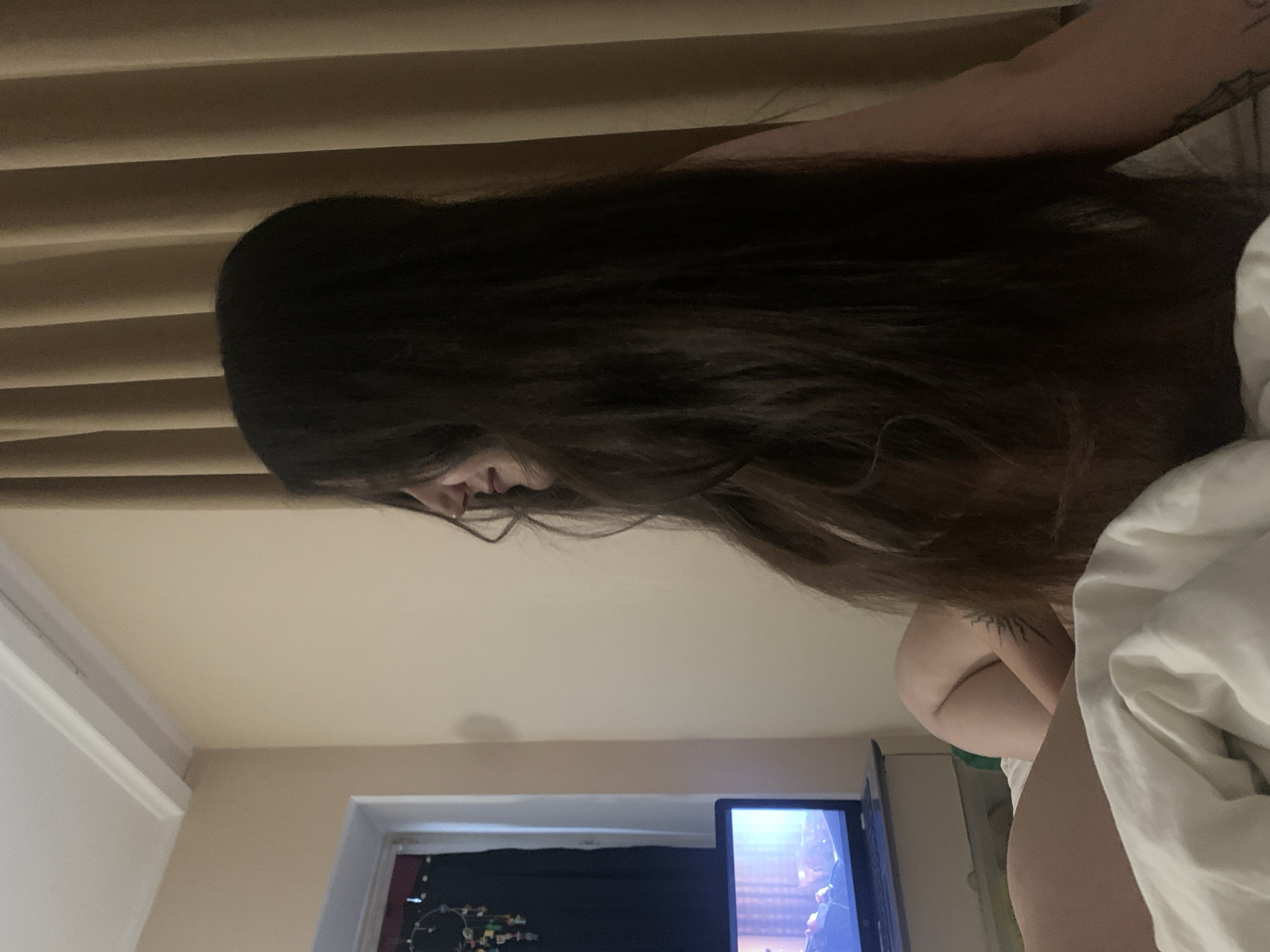 The image shows a woman with long dark hair lying in bed, facing away from the camera. She is looking at a laptop computer, which is open on the bed in front of her. The room is dimly lit, with beige curtains in the background and a window to the left. There is a dreamcatcher hanging on the window. The woman appears to be in a hotel room or a bedroom. The image does not provide any specific information about the location in the city of Сонячне, Ukraine.