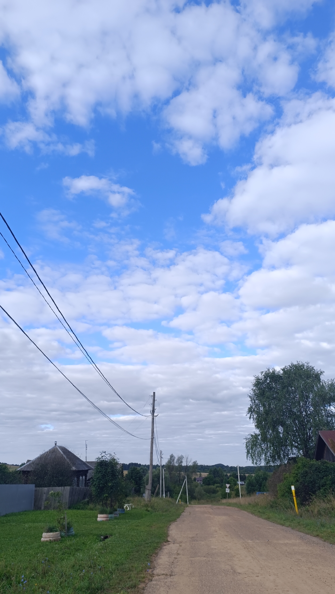 The image depicts a rural scene, likely in the city of Karsha, Russia, on a partly cloudy day. A dirt road runs through the center of the frame, leading into the distance. To the left of the road, there is a grassy area with a weathered wooden house and a grey fence. Several tire planters are visible on the lawn. A utility pole stands on the right side of the road, with power lines stretching across the upper portion of the image. In the background, rolling hills and scattered trees are visible. A large tree is situated to the right of the road, and part of a roof is visible behind it. The sky is a bright blue, with scattered white clouds. There are no people or animals present in the image.