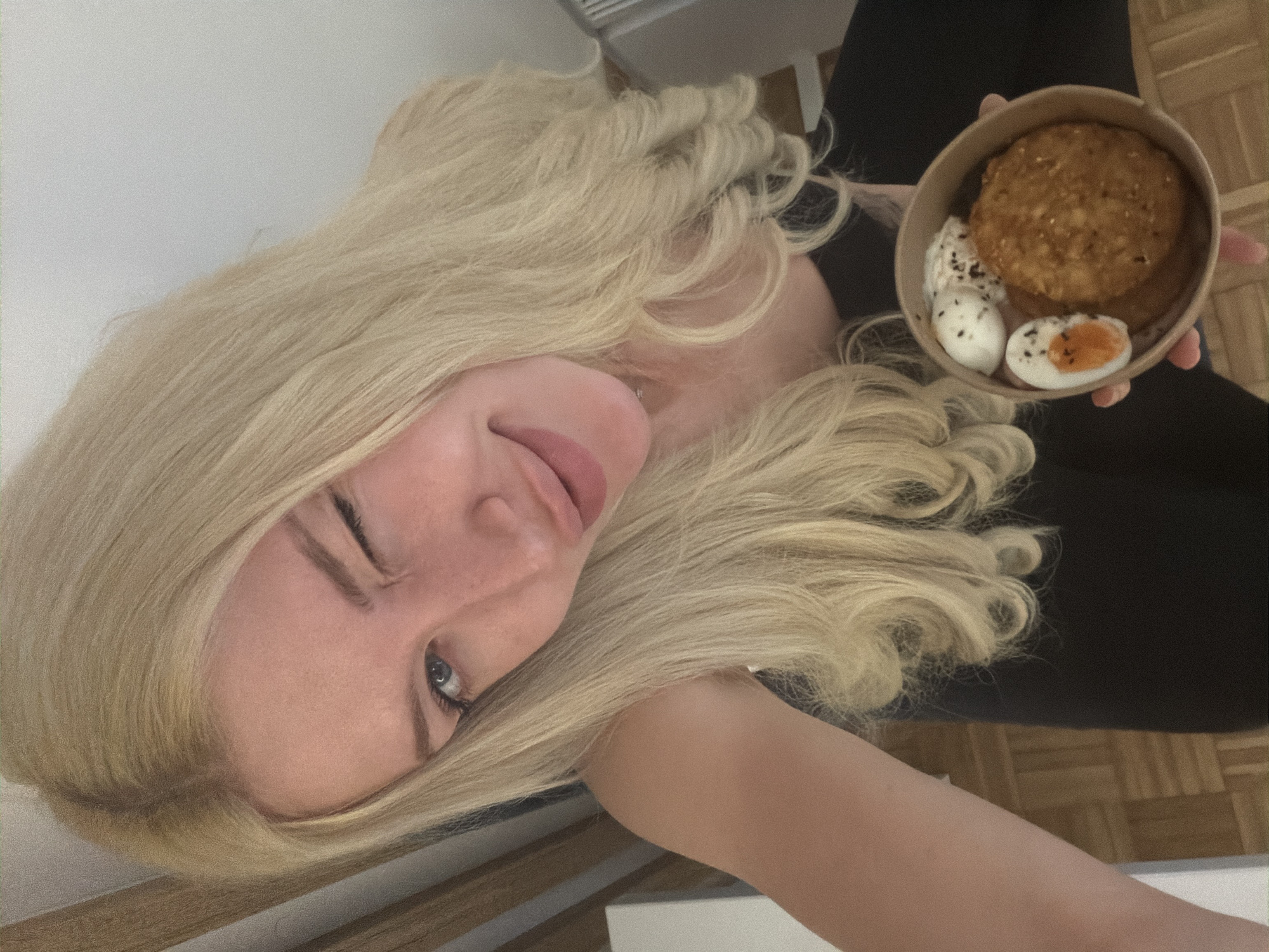 A young woman with blonde hair holds a bowl of food. The bowl contains what appears to be a burger and two halves of a boiled egg. The background is out of focus but seems to include a chair and a floor. There is no information to suggest that the image was taken in Belgrade, Serbia.