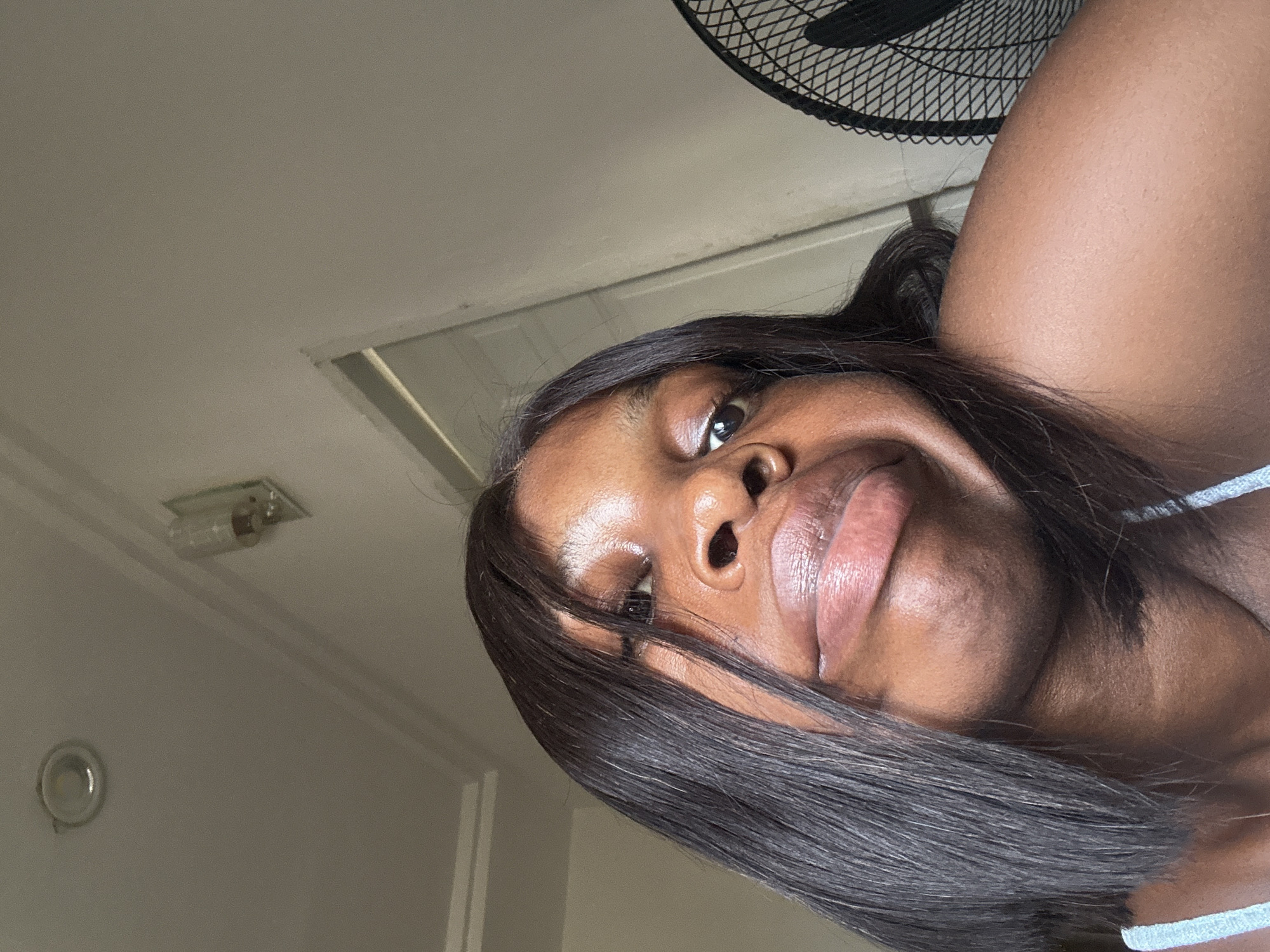 A close-up, tilted shot of a woman indoors. The woman has dark skin and dark hair, which is styled straight and reaches past her chin. Her features are prominent, and her lips are full. The background includes a ceiling with a light fixture, and a fan is partially visible in the upper right corner. Location details are not discernible to confirm the city of Abuja, Nigeria.