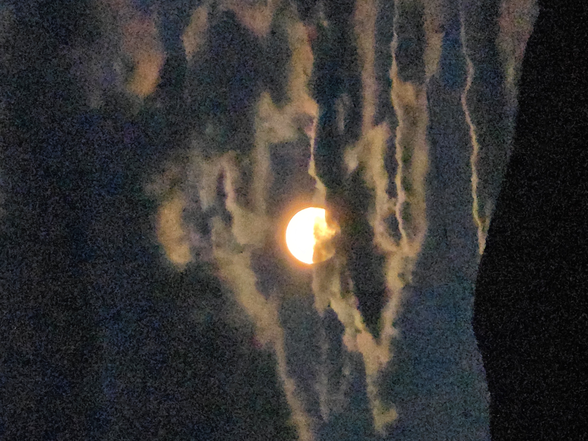 The image shows a partially obscured bright orb, likely the moon or sun, glowing with intense yellow and white light. It is positioned centrally in the frame, peeking through dark, textured clouds that are tinged with yellow and orange hues, suggesting twilight or dawn. The background is a deep, dark blue, creating a stark contrast with the illuminated orb and clouds. In the foreground, a dark, silhouetted landscape, possibly a mountain range or hilly terrain, occupies the bottom portion of the image. The overall impression is one of a dramatic celestial event against a dimly lit natural backdrop. The location in Magnitka, Russia, is a geographical detail not directly discernible from the visual content of the image itself.