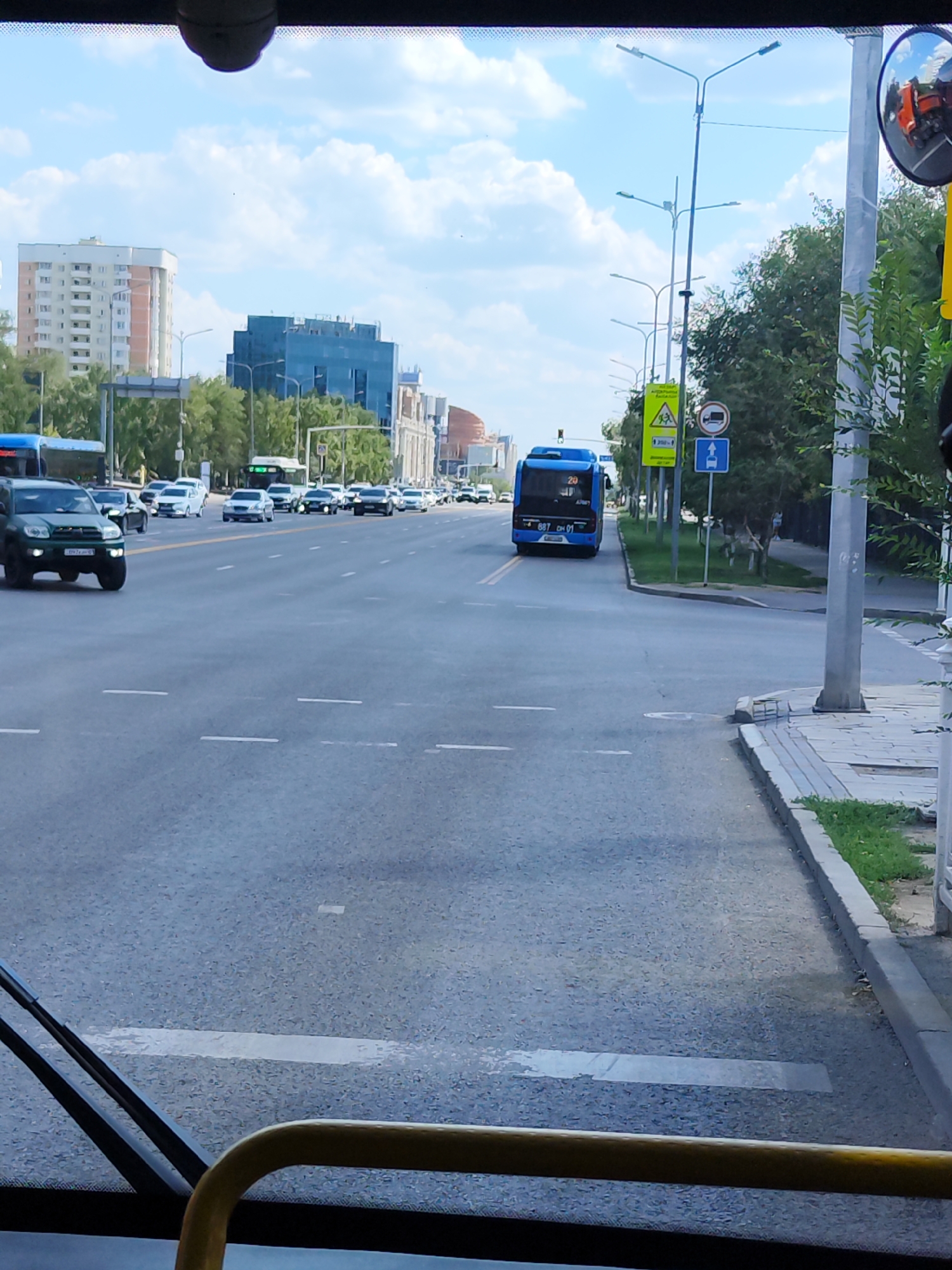 The image shows a street scene in Astana, Kazakhstan, on a bright, cloudy day. A blue bus with the number 20 on its front is traveling down the road. In front of the bus, a dark green SUV is also driving on the road. Several other vehicles, including white cars and a white bus, are visible in the distance. Tall buildings and trees line the street. A traffic light is visible, as are various traffic signs. A pedestrian crossing is marked on the road. The photo is taken from inside a vehicle, likely a bus, as indicated by the yellow railing and windshield wipers in the foreground. A reflection of an orange vehicle can be seen in a side mirror on the right side of the image.