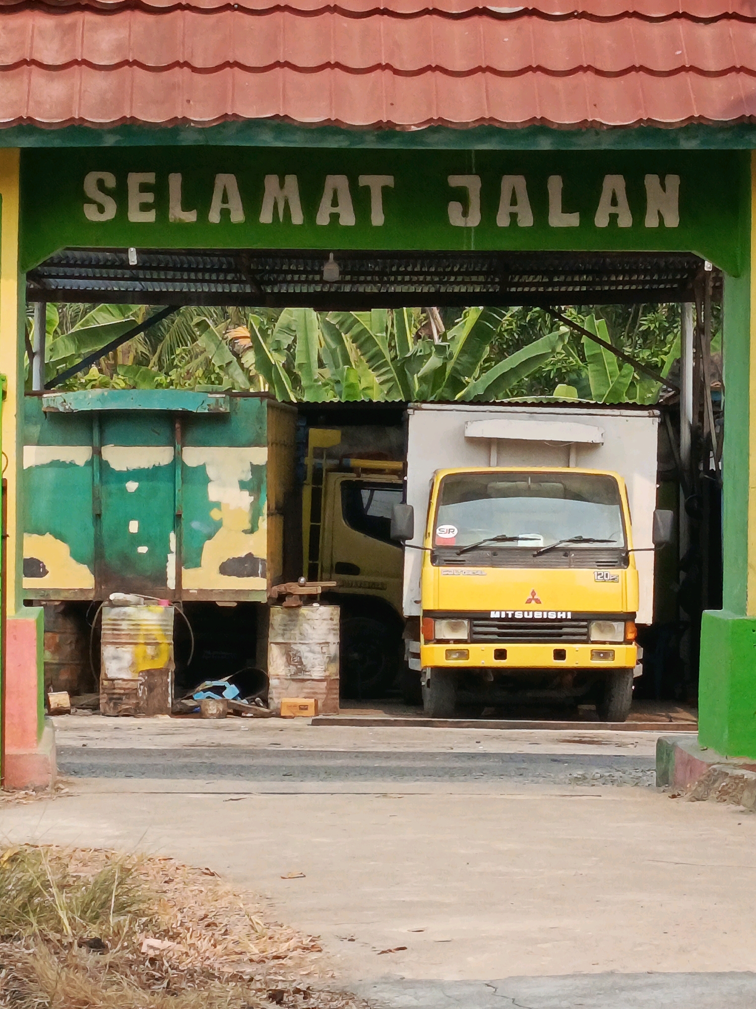 In Karya Dadi, Indonesia, a yellow Mitsubishi truck is parked under a covered structure. The truck has a white cargo box and is branded with "MITSUBISHI" and "120". Above the truck, a green sign with white lettering reads "SELAMAT JALAN," which translates to "Have a good trip" in Indonesian. To the left of the truck, a large green and yellow container is supported by several metal barrels. The background reveals lush green banana trees. The ground is paved with concrete and dirt.