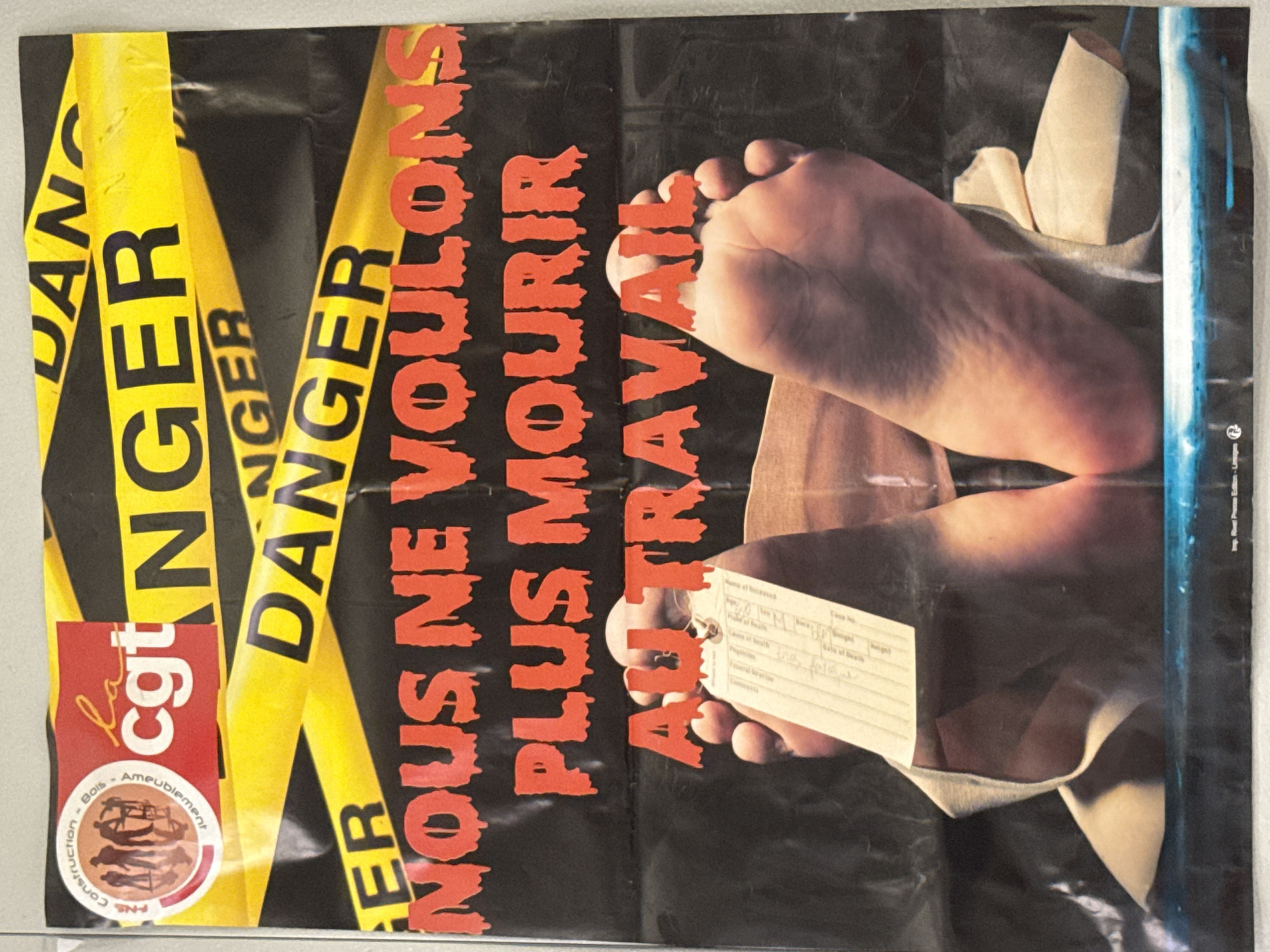 This media file is a poster with a dark background, featuring yellow "DANGER" tape. In the foreground, a close-up shot focuses on a human foot and lower leg, appearing to be bandaged and slightly injured. A white tag with handwritten text is attached to the leg, possibly indicating identification or cause of death, with fields like "Name of deceased," "Age," "Cause of death," and "Date of death" visible.
The poster displays bold, red, blood-like text in French that reads "NOUS NE VOULONS PLUS MOURIR AU TRAVAIL," translating to "WE DO NOT WANT TO DIE AT WORK." This strongly suggests the poster is a protest or awareness campaign against workplace fatalities or dangerous working conditions.
To the left of the foot, a circular logo is visible, containing the text "CGT," which is a major French trade union confederation, and further text indicating "Construction Bois Ameublement" (Construction Wood Furnishing). This reinforces the context of labor rights and safety in specific industries.
The overall tone of the poster is stark and impactful, aiming to evoke a sense of urgency and concern regarding worker safety. The use of "DANGER" tape and the graphic imagery of an injured foot contribute to this serious message. The poster appears to be printed, with a small imprint at the bottom right reading "Imp. Rivel Presse Edition - Lisages," which likely refers to the printing company and location in France. The lighting is artificial and focused on the subjects, creating a dramatic effect. There are no specific cues about the time of day or weather, as it is an indoor or studio-shot image for a poster.