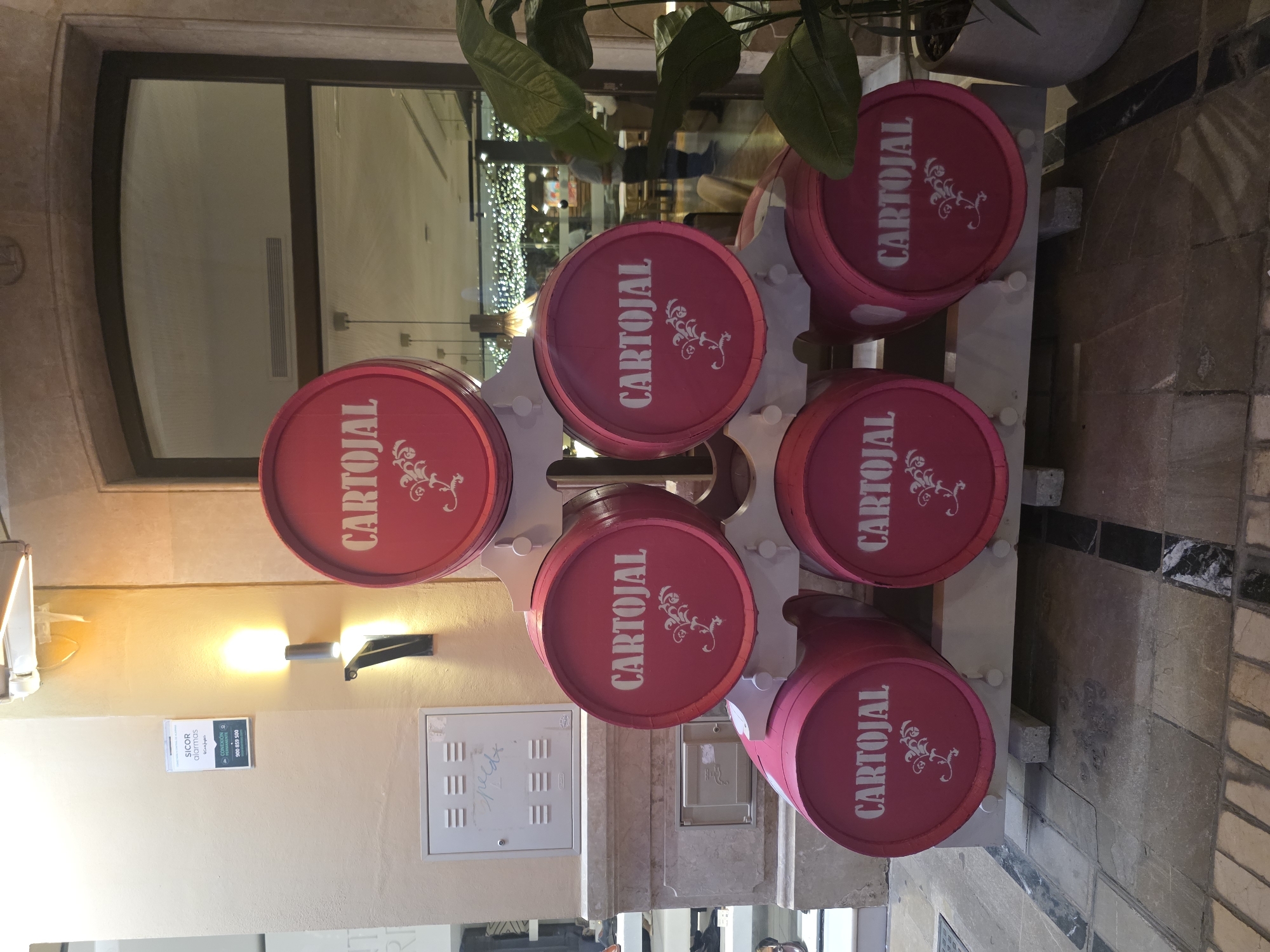 The image shows a decorative display of six bright pink barrels with "CARTOJAL" written in white script on each. The barrels are arranged in a tiered pyramid shape on a white wooden stand. The bottom row has three barrels, two of which are fully visible, and the top row has one barrel. The middle row has two barrels, both fully visible. The display is located outdoors, in what appears to be a pedestrian area with stone paving. In the background, there is a building with a large arched window reflecting an interior with tables, chairs, and decorative lights. A person in a blue shirt and dark pants is visible through the window. To the left of the barrels, there is a beige wall with a white electrical box and a white sign with blue text that reads "SICOR alarmas". There is also a metal lamp attached to the wall. To the right of the barrels, a large potted plant with green leaves is visible. The scene suggests a commercial establishment, possibly a restaurant or shop, in Málaga, Spain, based on the context provided.