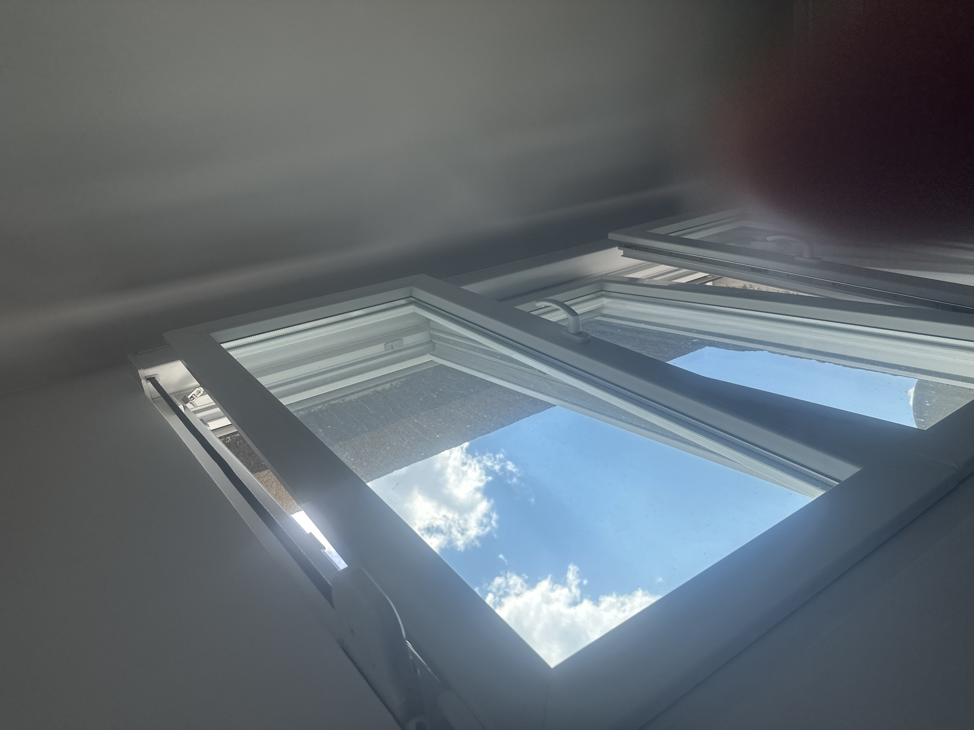 The media file shows an interior view of a white-framed window, possibly a skylight, angled upwards towards a bright blue sky filled with fluffy white clouds. The window appears to be open, allowing natural light to stream in. Sunlight glints off the window panes, creating a lens flare effect in the upper right portion of the image, where a reddish-brown circular blur suggests a person's hand or finger is present, partially obscuring the view.
The scene is set indoors, with the ceiling and walls of a room visible in the foreground, painted in a neutral, light color. The window frames are clean and modern. The perspective suggests the viewer is looking up from within a building. The bright sky and scattered clouds indicate a clear day, likely during daylight hours. There is no visible text in the image. The overall impression is one of a pleasant, sunny day seen from the comfort of an indoor space. The partial obstruction by a hand suggests a casual, perhaps personal, moment captured.