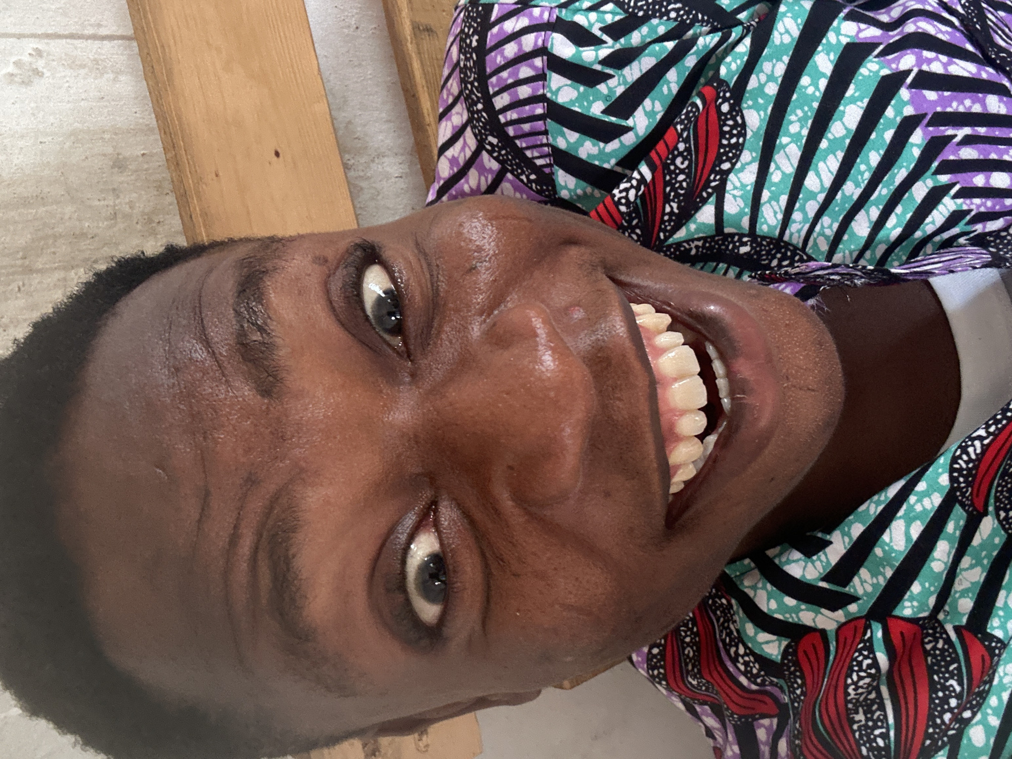 A close-up, rotated portrait depicts a person of African descent smiling broadly. Their skin appears dark and smooth, reflecting light. They have closely cropped, dark hair. Their eyes are open and grey, and their teeth are white and prominent. The person is wearing a colorful patterned garment with stripes of purple, black, turquoise, and red with white accents. In the background, a portion of a wooden structure is visible. The image's location is Unknown, Unknown.