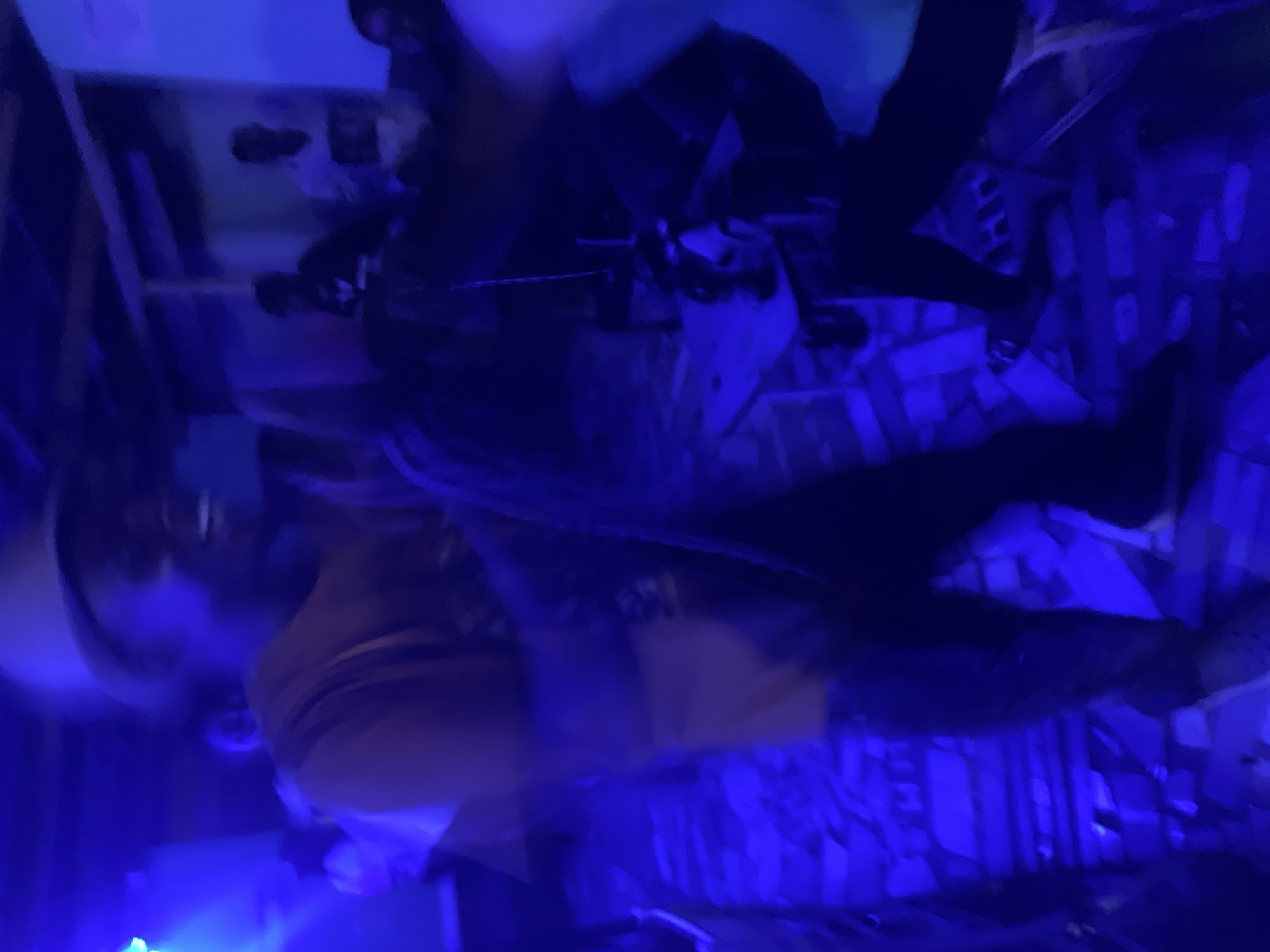 The image depicts an indoor social gathering, bathed in prominent blue light, located in New Stadium, Nigeria. A central figure, captured with significant motion blur, stands towards the left of the frame. This individual wears a light-colored hat (possibly beige or tan), a similarly colored short-sleeved top, and dark trousers. Their face is partially obscured by blur, but a clear smile with visible teeth is apparent, indicating a moment of laughter or speech. Their right arm is extended downwards and blurred, while their left arm is less distinct. Dark footwear, possibly with visible perforations, is worn on a tiled floor that appears uneven or covered with scattered debris, giving it a fragmented appearance.
To the right of the central figure, a small table is visible, holding a dark bottle, a glass, and other indistinct items. A thin, light-colored cord or chain hangs vertically near the table. Further into the background, several other individuals are present. On the far right, two figures are seated or standing against a wall; one wears a dark top, and the other a light-colored shirt featuring a dark graphic design. Architectural elements, including wooden beams, are visible near the ceiling. A bright blue light source emanates from the upper left, casting a strong blue hue across the entire scene and contributing to the overall low-light, high-motion blur aesthetic. The environment suggests a casual venue characterized by dim, colored lighting and activity.