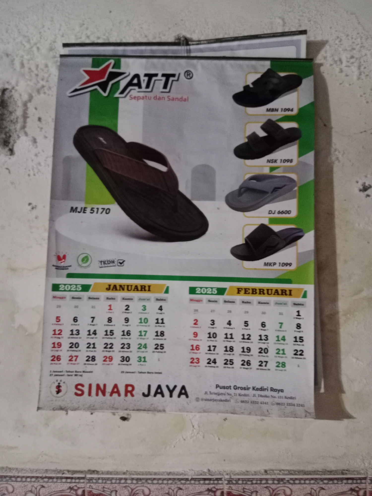 This is a close-up, eye-level, slightly angled photograph of a 2025 calendar featuring products from "ATT" brand footwear. The calendar displays January and February. The top portion of the calendar shows product images of various sandals with their model numbers: MJE 5170, MBN 1094, NSK 1098, DJ 6600, and MKP 1099. The calendar itself is mounted on a textured, off-white wall with visible stains and imperfections. Below the calendar, a business name "SINAR JAYA" is prominently displayed, along with contact information including an address: "Pusat Grosir Kediri Raya, Jl. Sriwijaya No. 71 Kediri, Jl. Dhono No. 131 Kediri," and phone numbers. The calendar has a slight curl at the bottom edge. The lighting appears to be ambient. There are no people or animals visible in the image. The overall setting suggests an interior space, possibly a store or office. The reference to Kediri in the address indicates the geographical context.