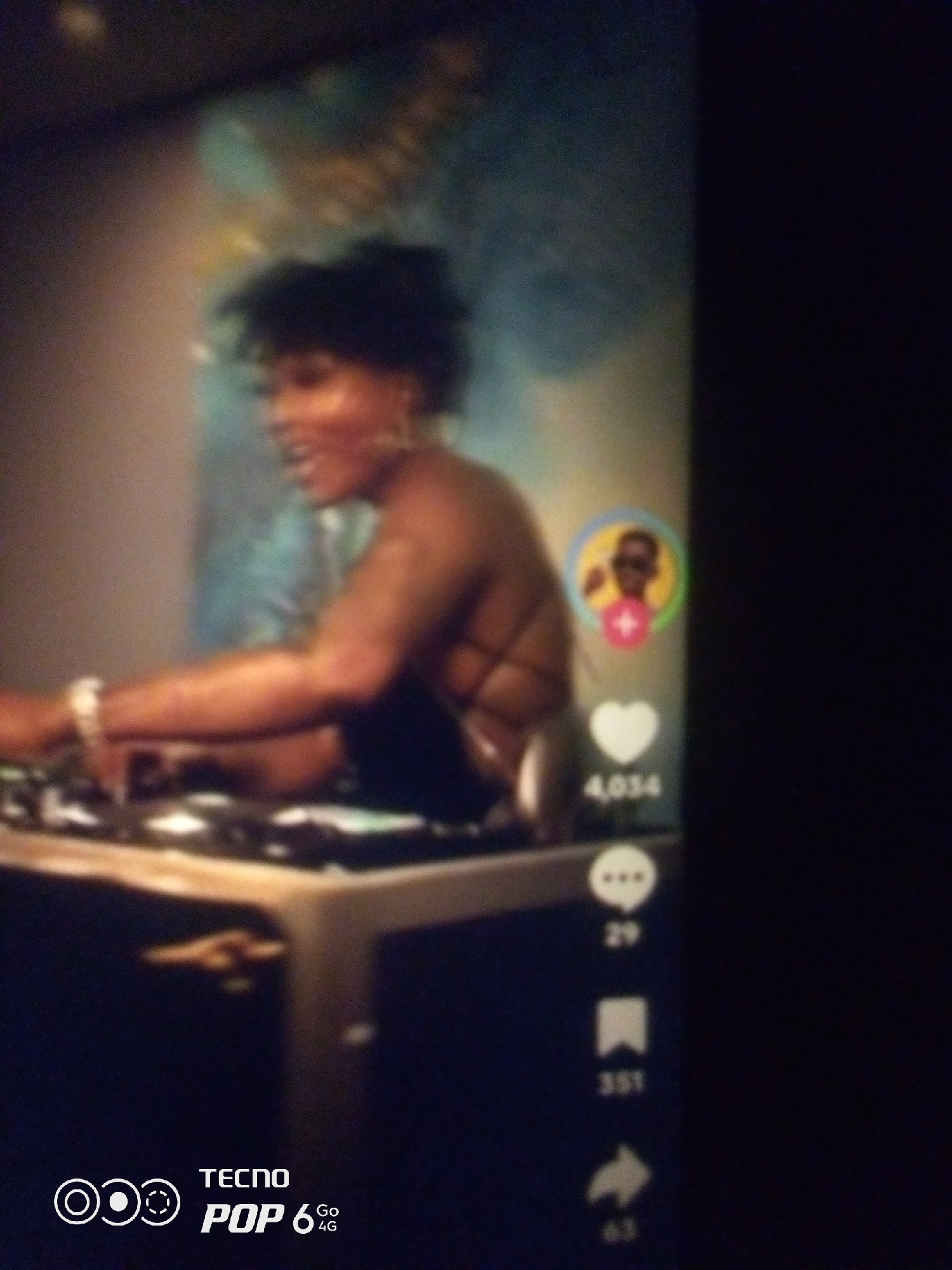 The image shows a person with dark, curly hair, wearing a dark top and a bracelet on their left wrist, sitting in front of a DJ setup. The person appears to be a woman with brown skin and is smiling. Behind the person is a blurry background with what looks like a painting or mural. On the right side of the image, there are social media icons: a profile picture within a circle with a plus sign, a heart icon with the number 4,034 below it, a speech bubble icon with the number 29 below it, a bookmark icon with the number 351 below it, and a share icon with the number 63 below it. At the bottom left corner, there is a watermark that says "TECNO POP 6 4G". The image appears to be a screenshot of a video or a picture taken from a screen. The scene is indoors, and the lighting is dim. There is no indication of the specific location within Howayi, Nigeria.