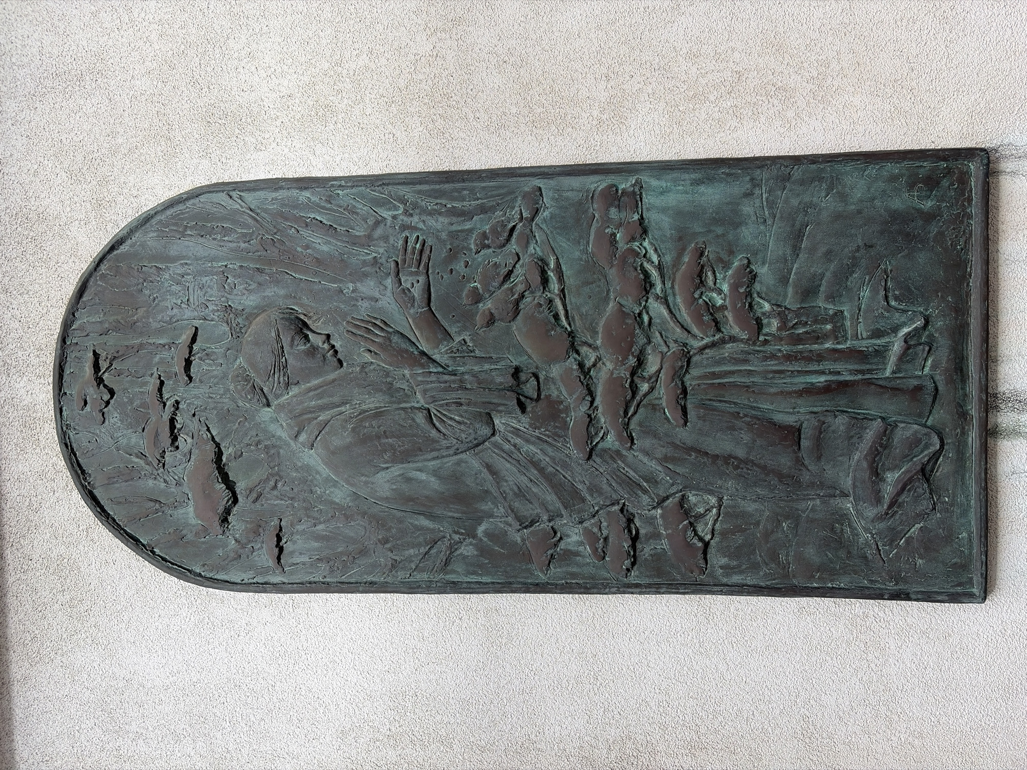 A vertical, arched bronze relief sculpture is mounted on a textured, off-white stucco wall. The sculpture depicts a robed figure, possibly a monk, kneeling or bowing with hands extended and open. The figure's head is bowed in prayer or supplication. Several small birds are perched on branches or in the air around the figure, and the relief also includes stylized trees or foliage. The bronze has a patina with areas of green oxidation. The bottom right corner of the relief shows a small, incised monogram. The overall scene evokes a sense of serenity and communion with nature. The specific location of this artwork within the city of Unknown, Unknown is not identifiable from the image.