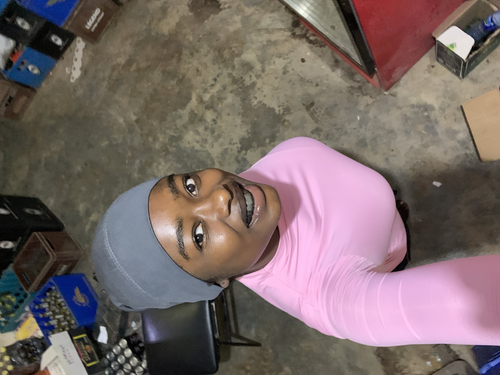 A young woman wearing a light pink shirt and a gray head covering is looking up at the camera. The environment appears to be a storage area with multiple boxes and crates scattered on the floor. A red refrigerator is visible in the background, and a black chair is located near the woman. The location is assumed to be New Stadium, Nigeria.