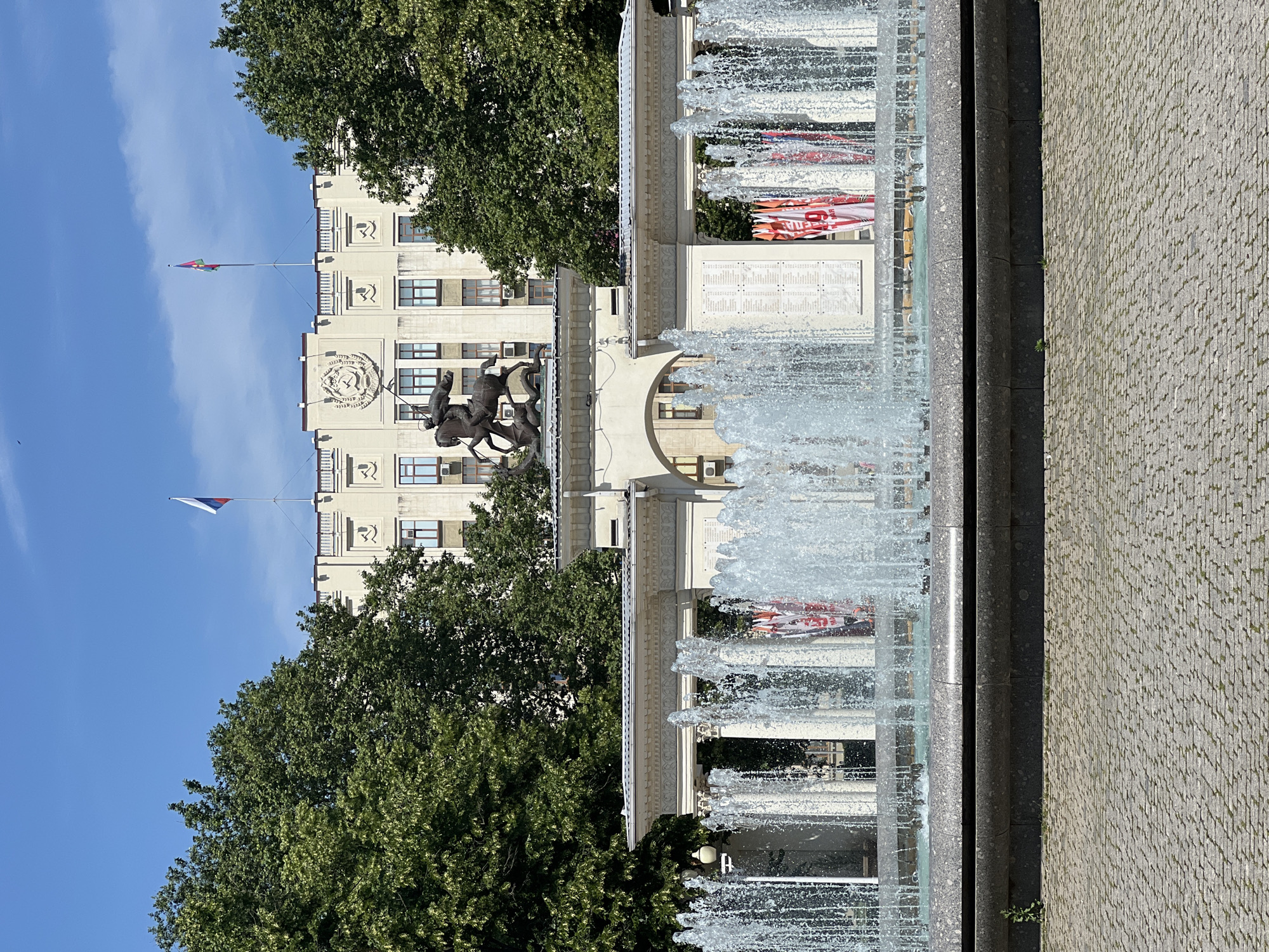 The image depicts a view of the Krasnodar administration building from across a fountain. The building, a large, light-colored structure with multiple windows and decorative elements, is partially obscured by lush green trees on either side. Two flags fly from the rooftop. In front of the building is a colonnaded structure with an arched entryway. Mounted above this structure is a bronze equestrian statue of Saint George slaying a dragon. The foreground is dominated by a multi-tiered fountain with water spraying upwards. The ground in front of the fountain is paved with cobblestones. Several red and white banners are visible behind the fountain, attached to the colonnaded structure. The sky is clear and blue.