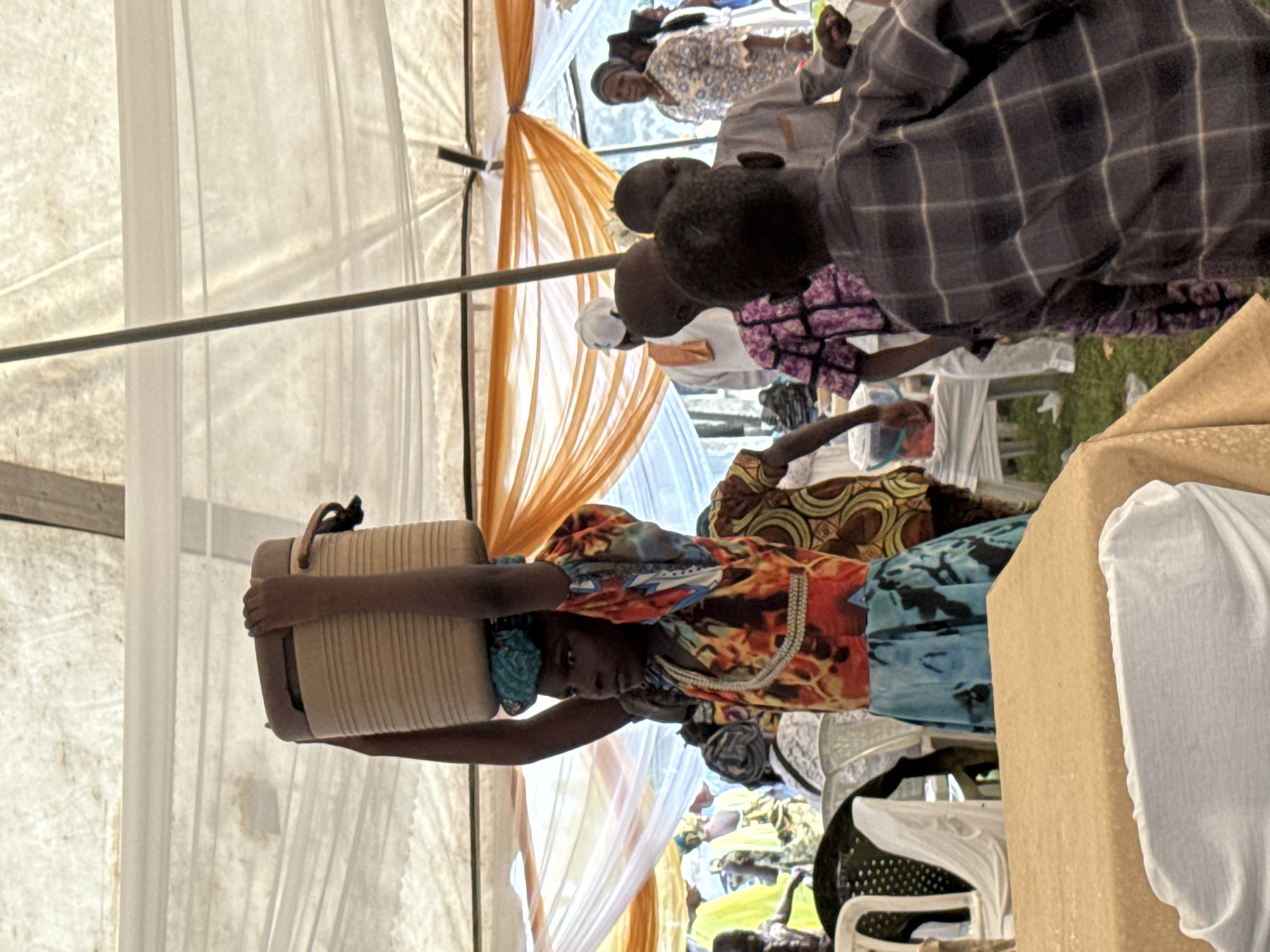 In a brightly lit outdoor gathering under a white tent with orange draping, a young person is the central focus, balancing a large, beige, ribbed container on their head. Their arms are raised to steady the load. They are wearing a vibrant, patterned shirt with yellow, red, and blue hues, and a blue and green patterned skirt. A blue cloth is tied around their head.
Around them, other individuals are visible, suggesting a communal event. In the background, a woman in a blue and white patterned dress stands to the right, and other people in colorful attire are scattered throughout the scene. Several white chairs and a table covered in a tan cloth are also present. The overall environment appears to be a festive or ceremonial gathering, potentially an outdoor event in a Nigerian city like Badagry, given the cultural clothing and tent structure. The image captures a moment of activity and community participation.
