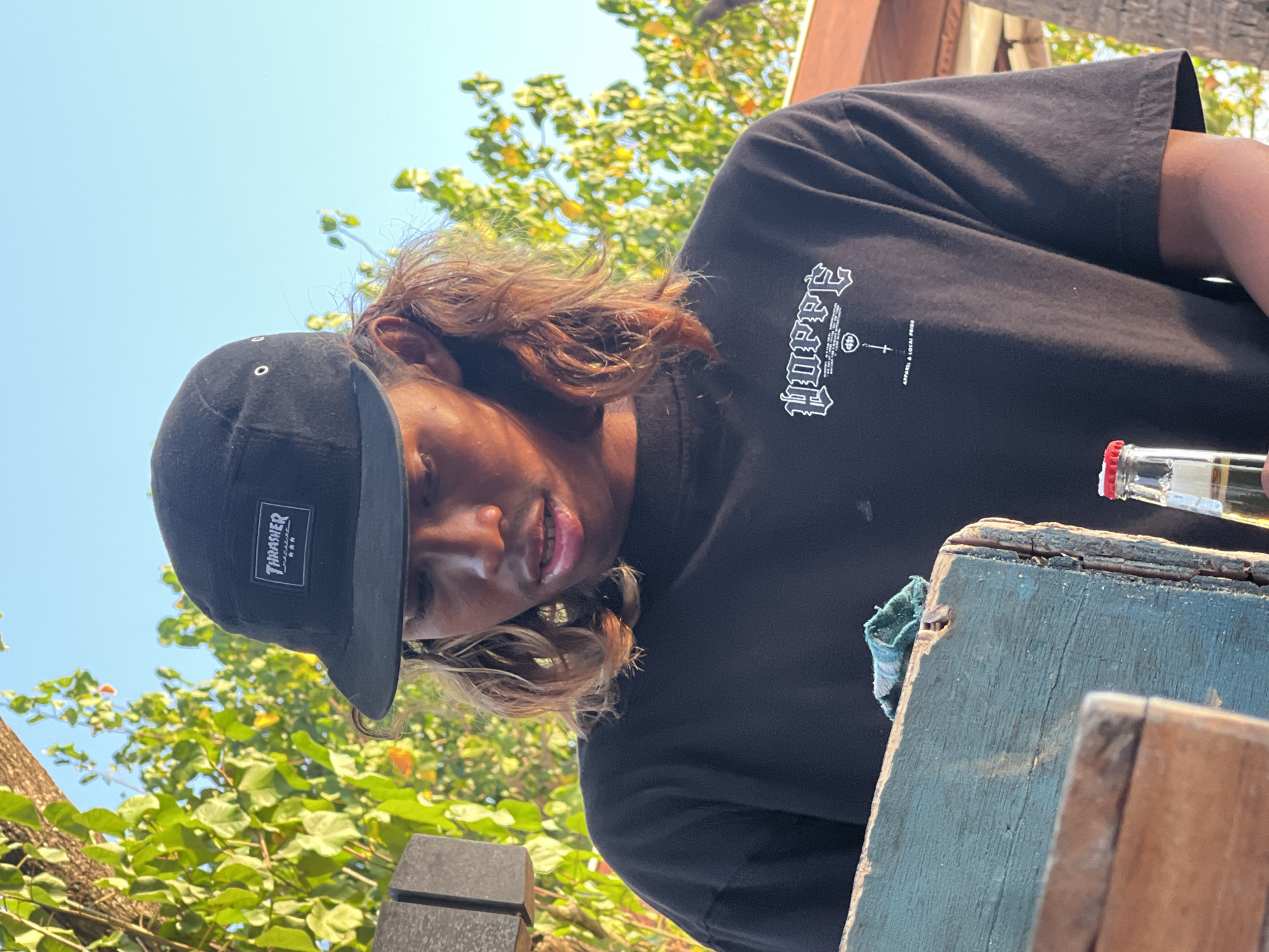 A young man wearing a black t-shirt and a black baseball cap with the "Thrasher" logo is standing outdoors. He has long, light brown hair and is looking down. In the foreground, a light blue wooden box is visible, with a small piece of blue and white fabric on top. To the right of the box, a glass bottle with a red cap is partially visible. The background consists of green foliage and a clear blue sky. The lighting suggests it is daytime, likely during the late afternoon given the warm tones. The overall impression is of a casual, outdoor setting.